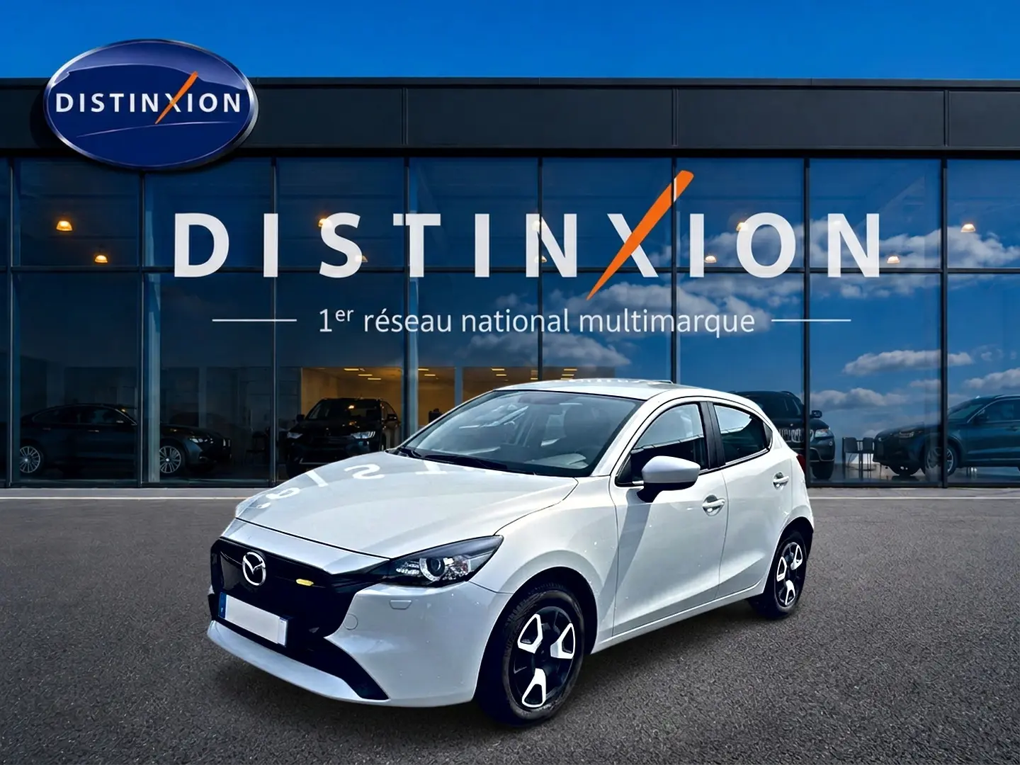 Vue 3/4 avant droite d'une Mazda2 blanche 2024 avec jantes noires et calandre noire devant un showroom Distinxion.