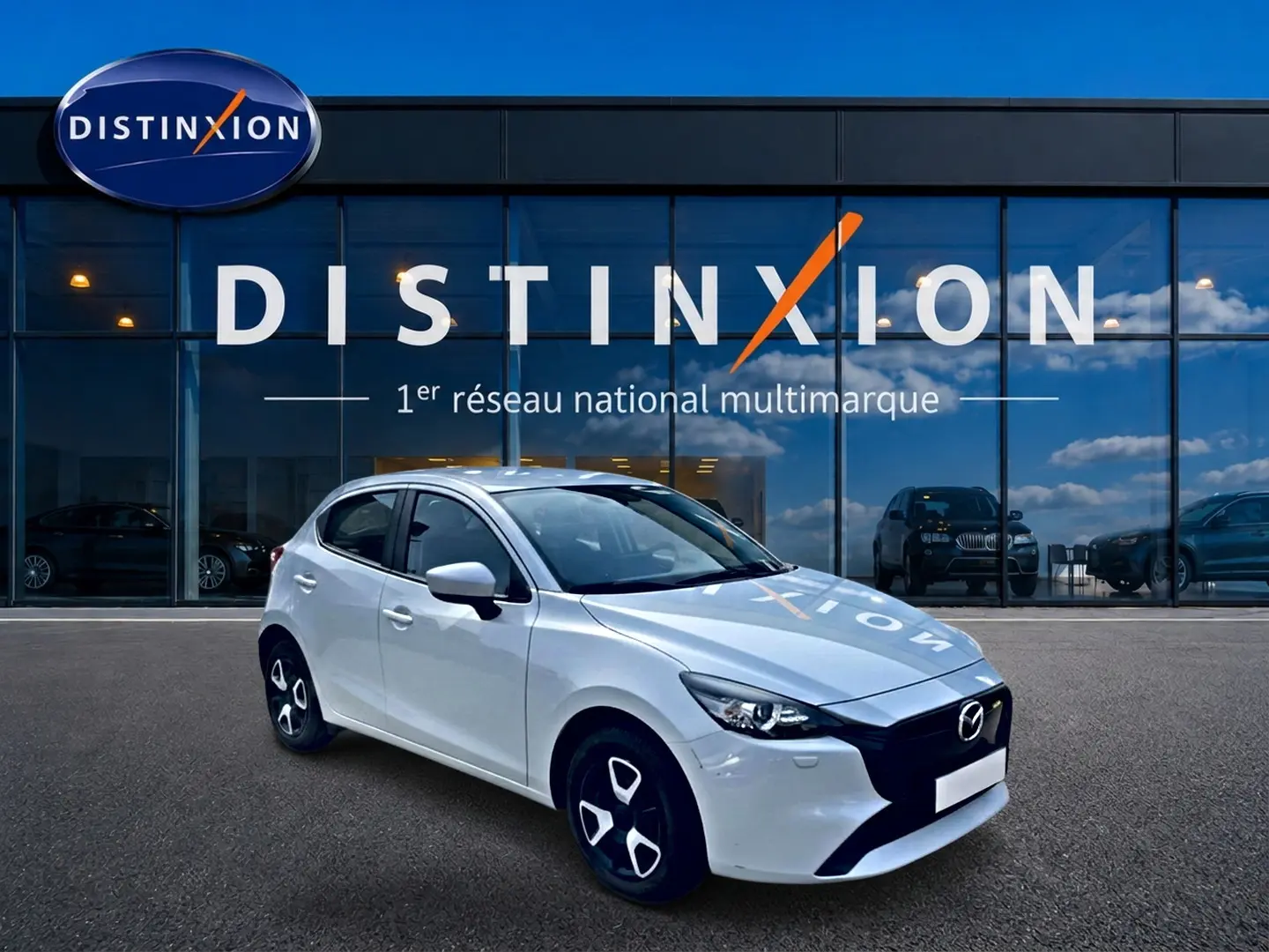 Vue 3/4 avant droite d'une Mazda2 blanche 2024 avec jantes noires et calandre noire devant un showroom Distinxion.
