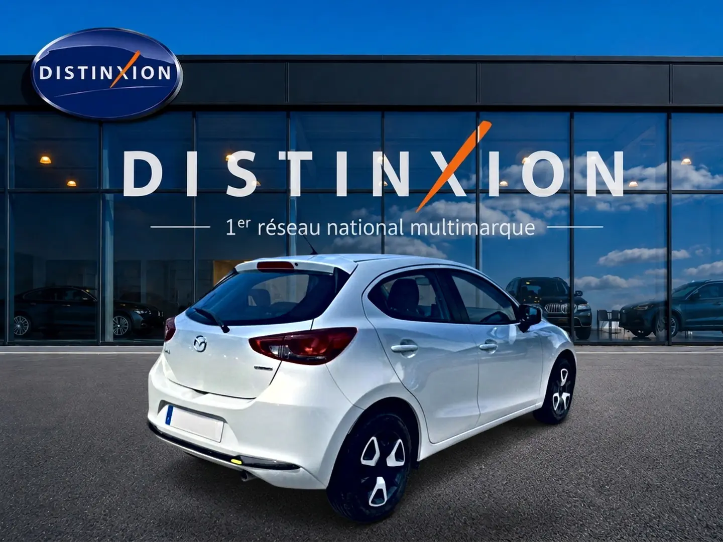 Vue 3/4 arrière droite d'une Mazda2 blanche 2024 garée devant une vitrine avec logo Distinxion.