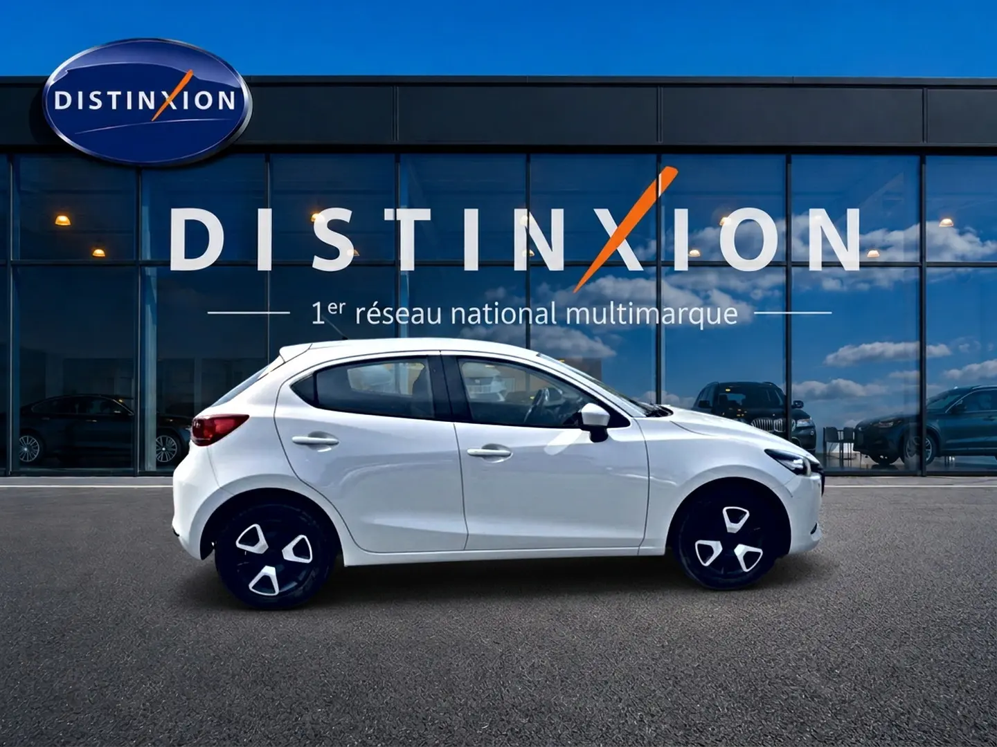 Vue latérale droite d'une Mazda2 blanche 2024 garée devant une vitrine avec logo Distinxion.