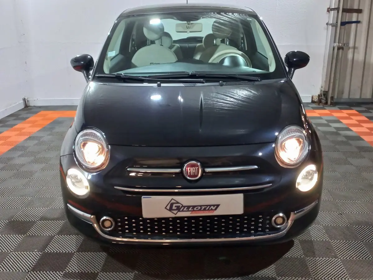 Vue de face d'une FIAT 500 noire avec phares allumés et intérieur clair visible au travers du pare-brise.