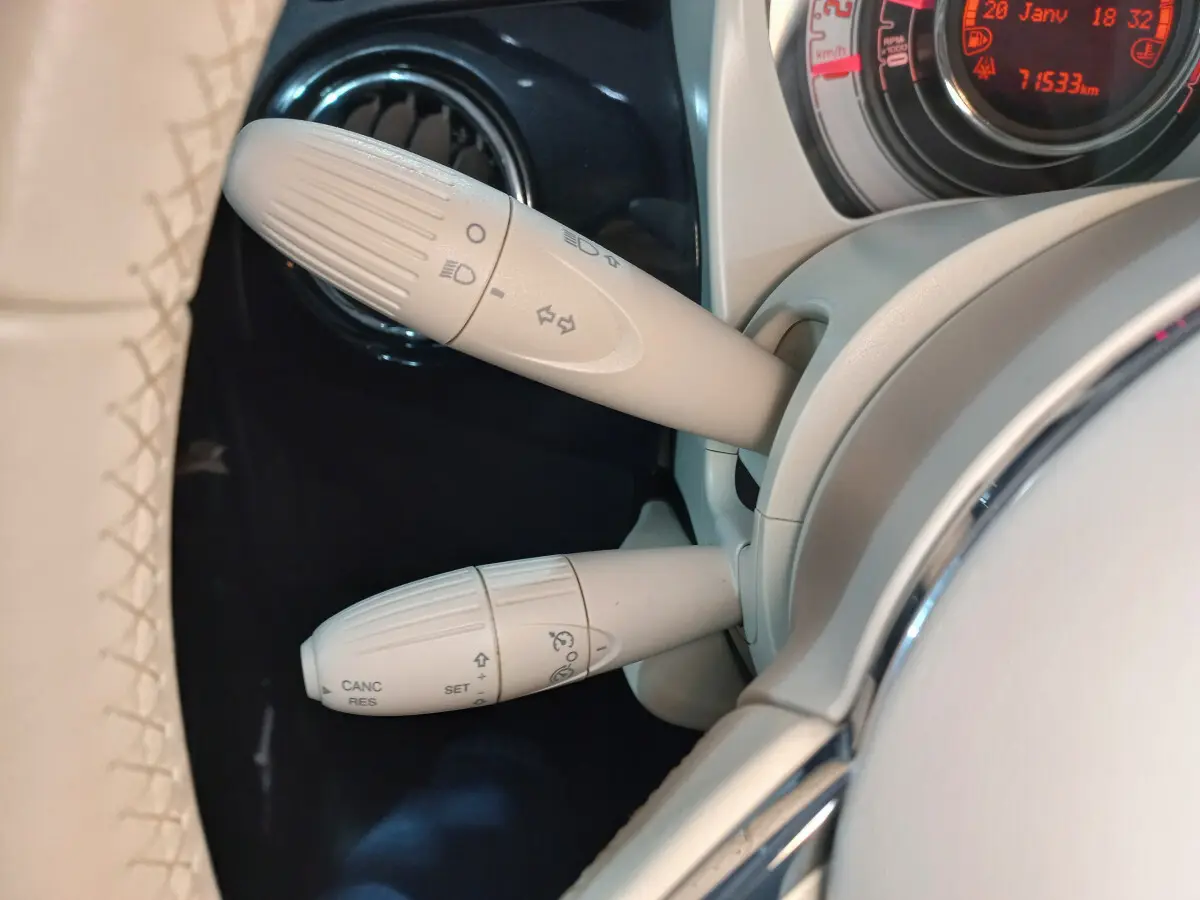 Gros plan sur les commandes beiges du volant cuir de la Fiat 500 Lounge noire, avec tableau de bord digital rouge en arrière-plan.