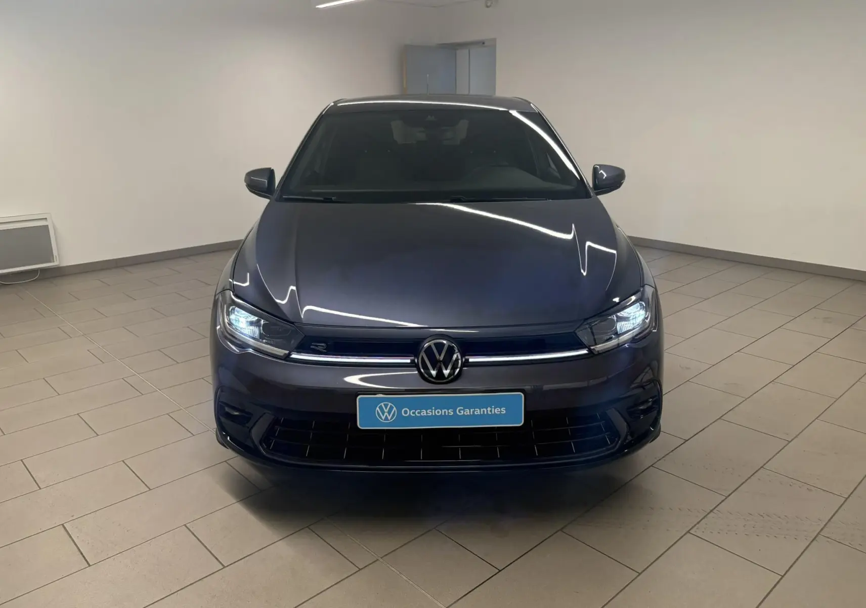 Volkswagen Polo 1.0 TSI R-Line gris cendré vue de face en intérieur avec phares allumés et logo VW visible.