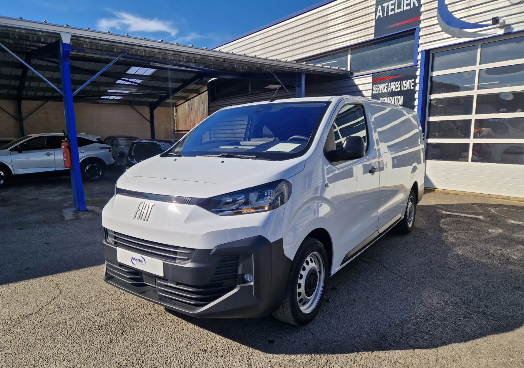Fourgon FIAT Scudo blanc en 3/4 avant droit, avec pare-chocs noir et calandre moderne, stationné devant un atelier.