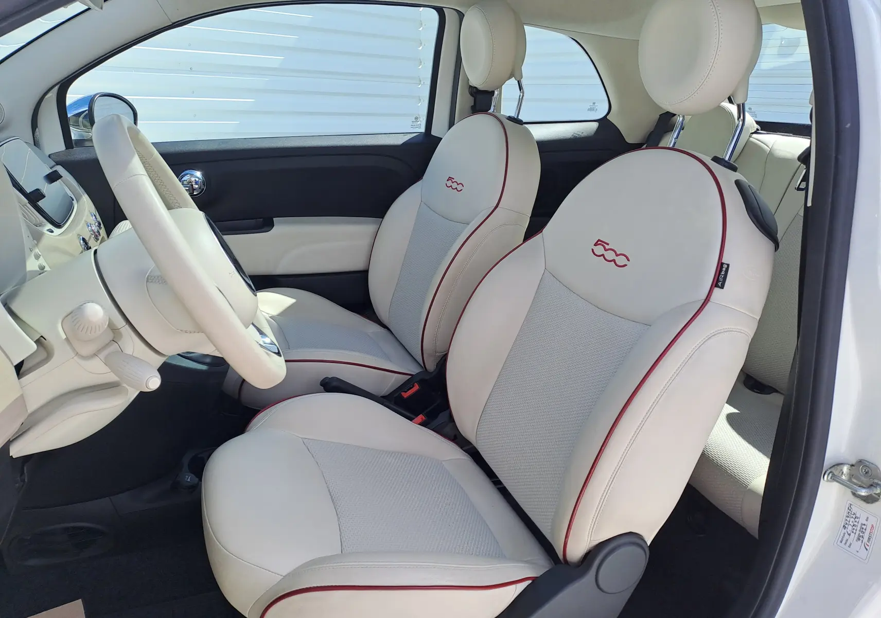 Intérieur blanc crème de la Fiat 500 1.0 Hybrid 2022, vue côté gauche sur les sièges avant avec logo 500 rouge.