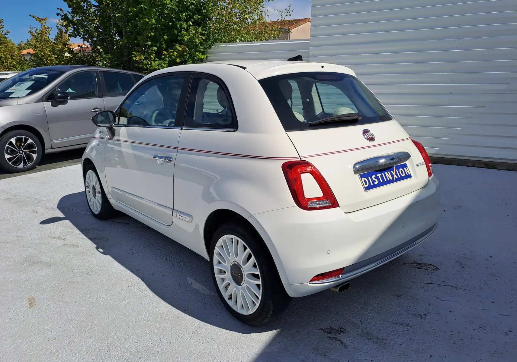 Fiat 500 blanche vue 3/4 arrière droit, avec jantes blanches et bande rouge décorative latérale.