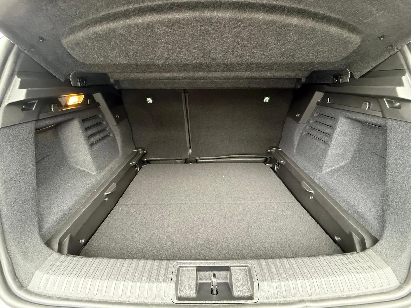 Coffre ouvert vu de l'arrière, intérieur spacieux et gris foncé, éclairage latéral à gauche visible.