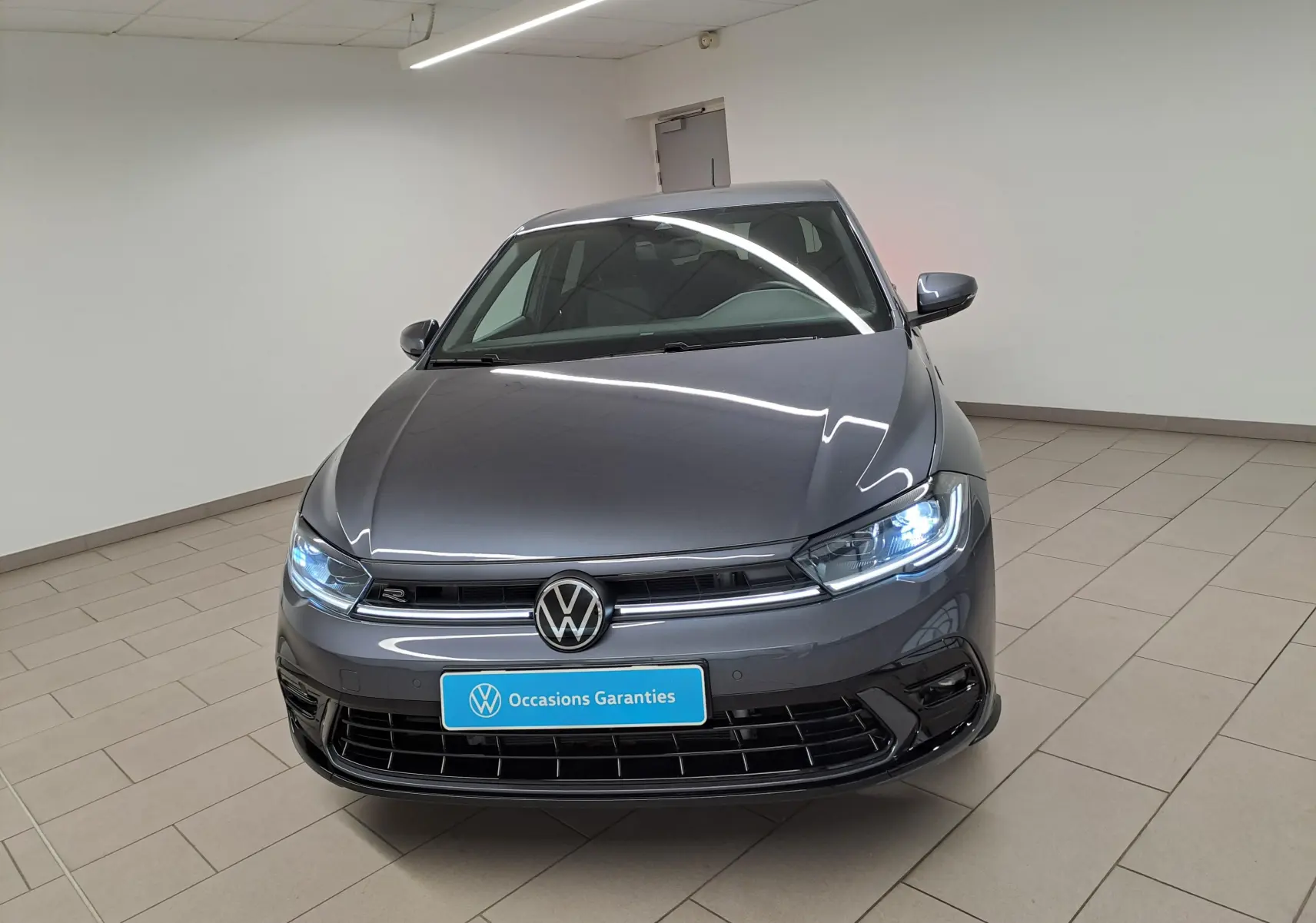 Vue avant d'une Volkswagen Polo 1.0 TSI R-Line gris cendré avec phares allumés dans un showroom.