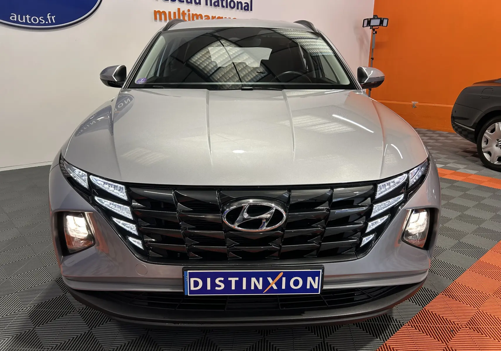 Vue avant d'un Hyundai Tucson hybride gris Shimmering Metal avec calandre noire et feux LED allumés en intérieur showroom.