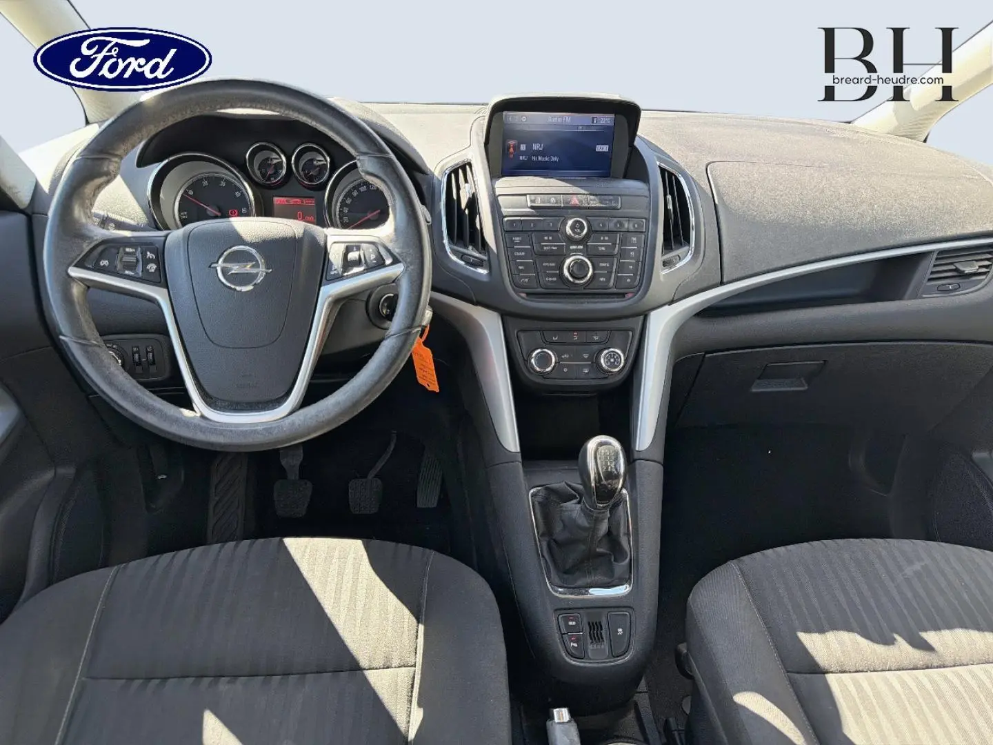 Vue intérieure avant de l’Opel Zafira Tourer 2013, tableau de bord et volant avec logo Opel, sellerie tissu gris.