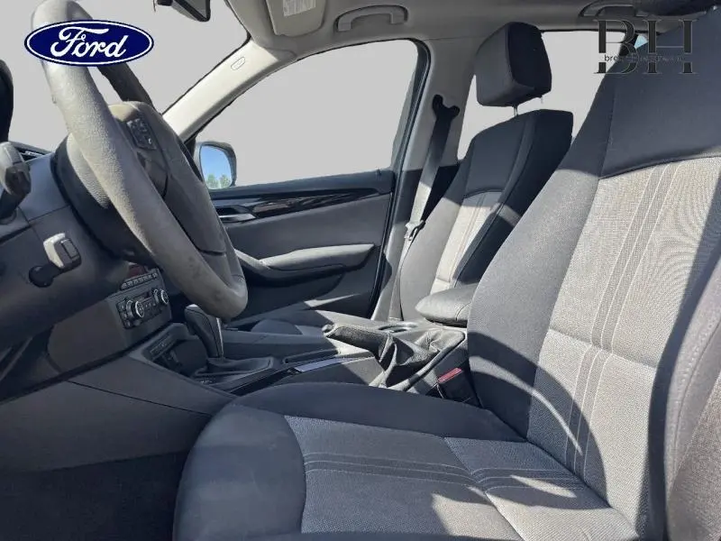 Vue intérieure côté conducteur du BMW X1 gris 2011, montrant les sièges avant et le volant multifonction en cuir.