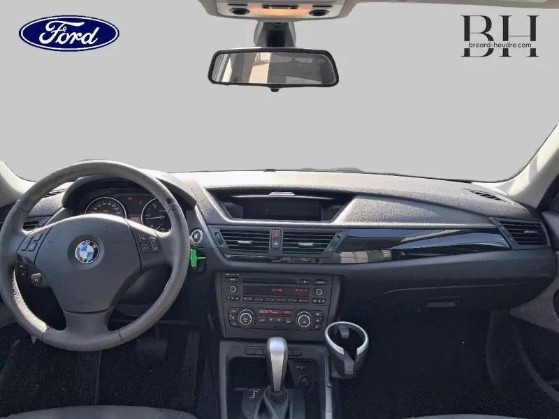 Vue intérieure avant du BMW X1 gris 2011 montrant le tableau de bord, volant cuir multifonction et console centrale avec boîte auto.
