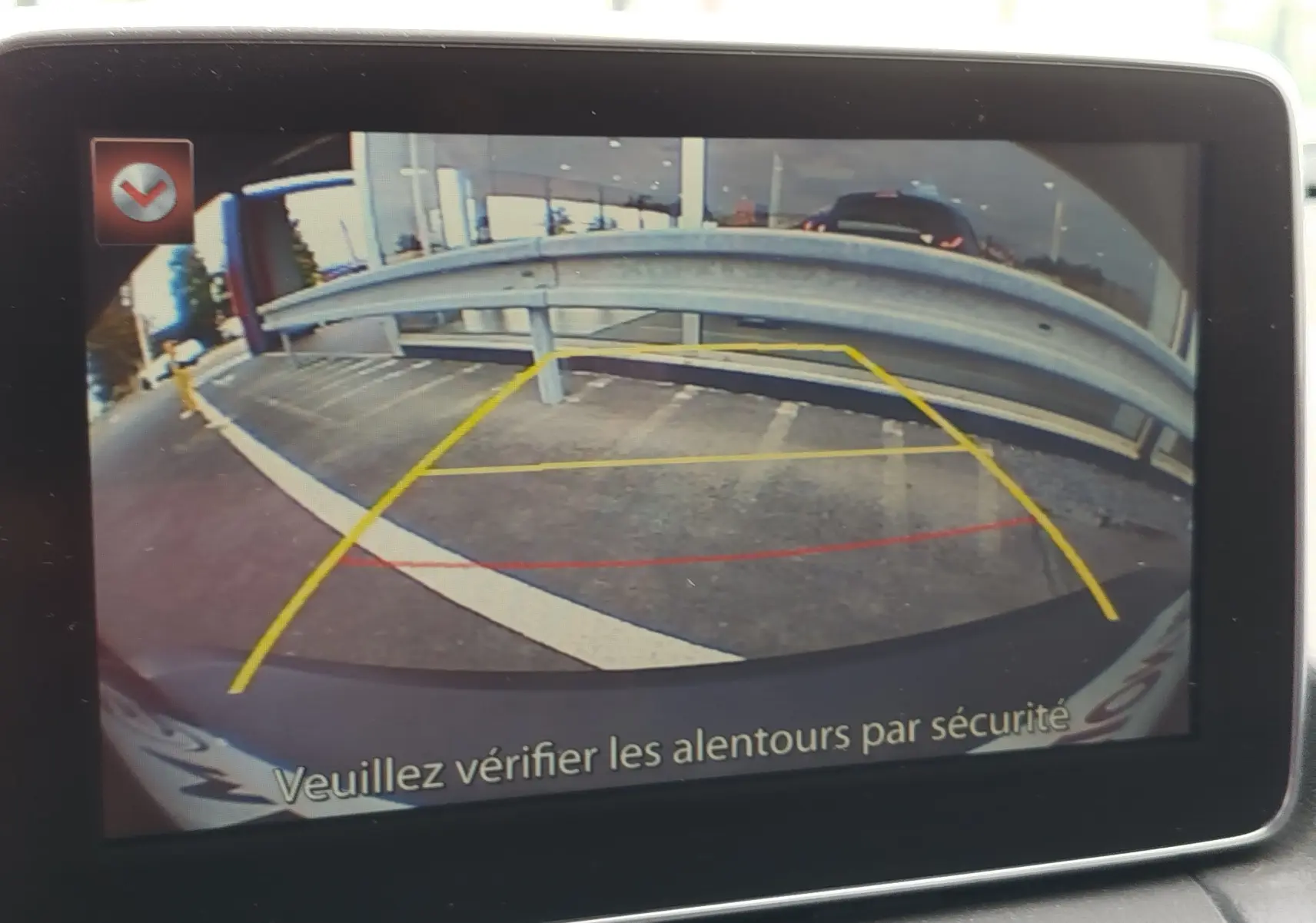 Vue caméra de recul du Mazda CX-3 bleu Dynamic Blue Mica, montrant la zone de stationnement et la barrière métallique arrière.