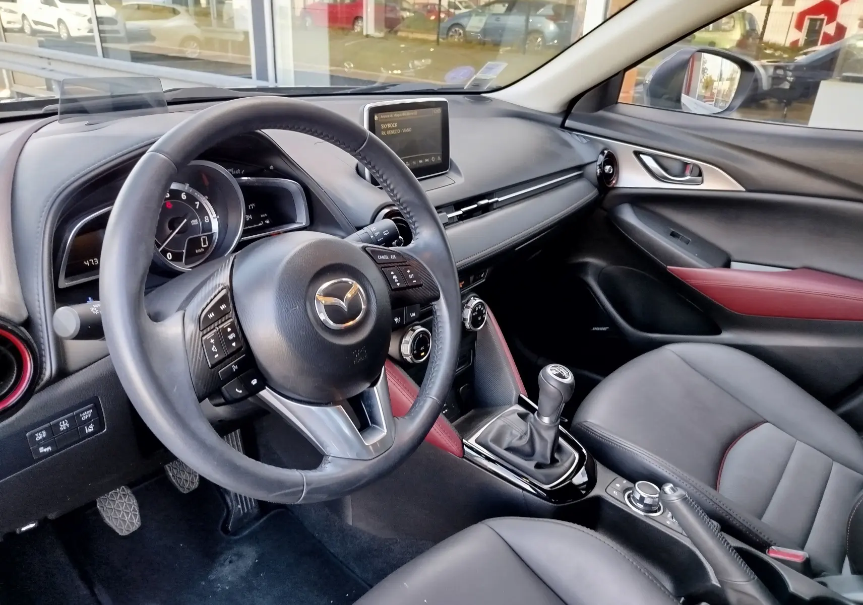 Intérieur du Mazda CX-3 2016 vu côté conducteur, volant cuir, sièges noirs avec surpiqûres rouges et levier de vitesse manuel.