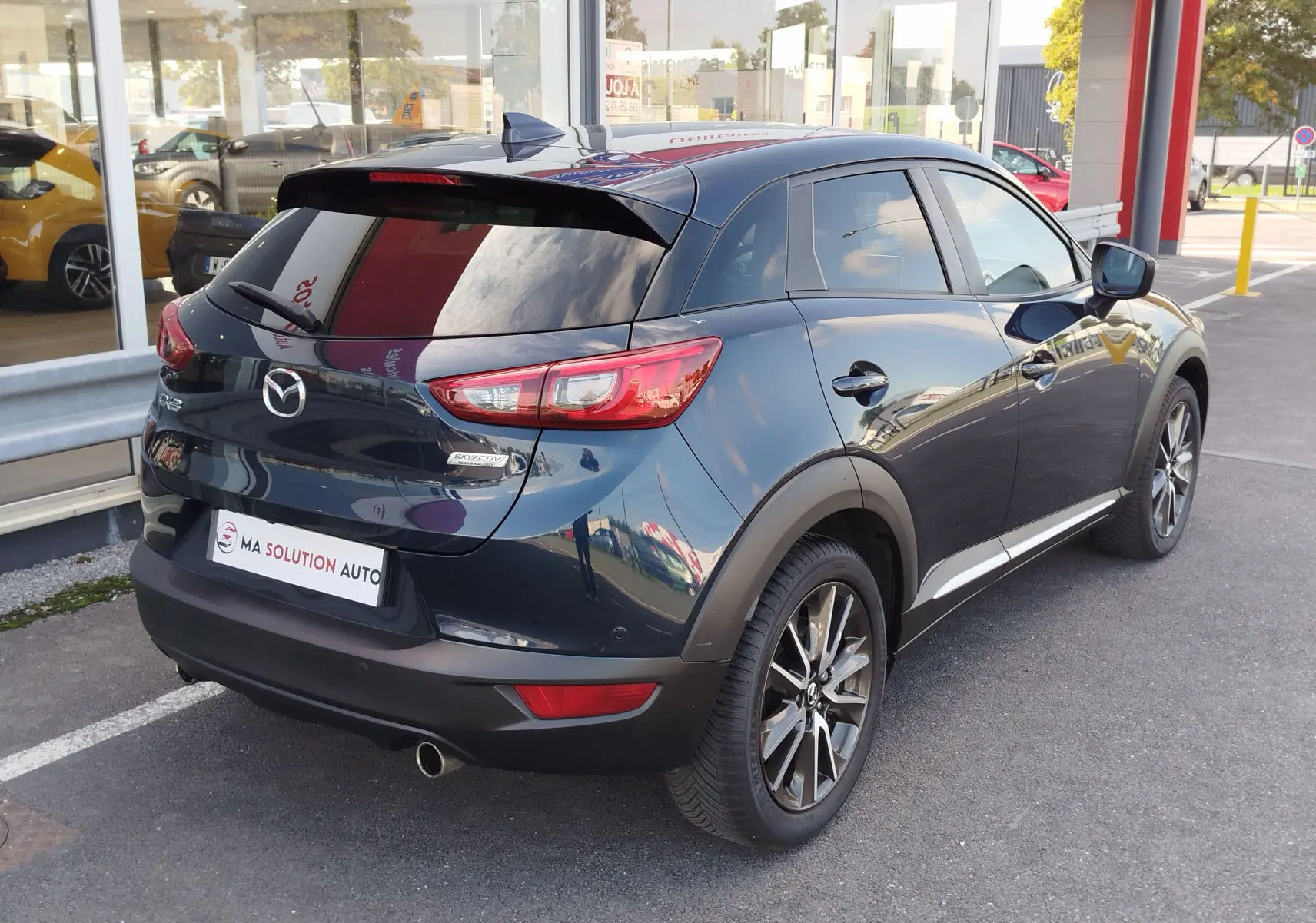 Vue 3/4 arrière droite d'un Mazda CX-3 bleu foncé avec jantes alliage et vitres teintées, stationné devant un showroom.