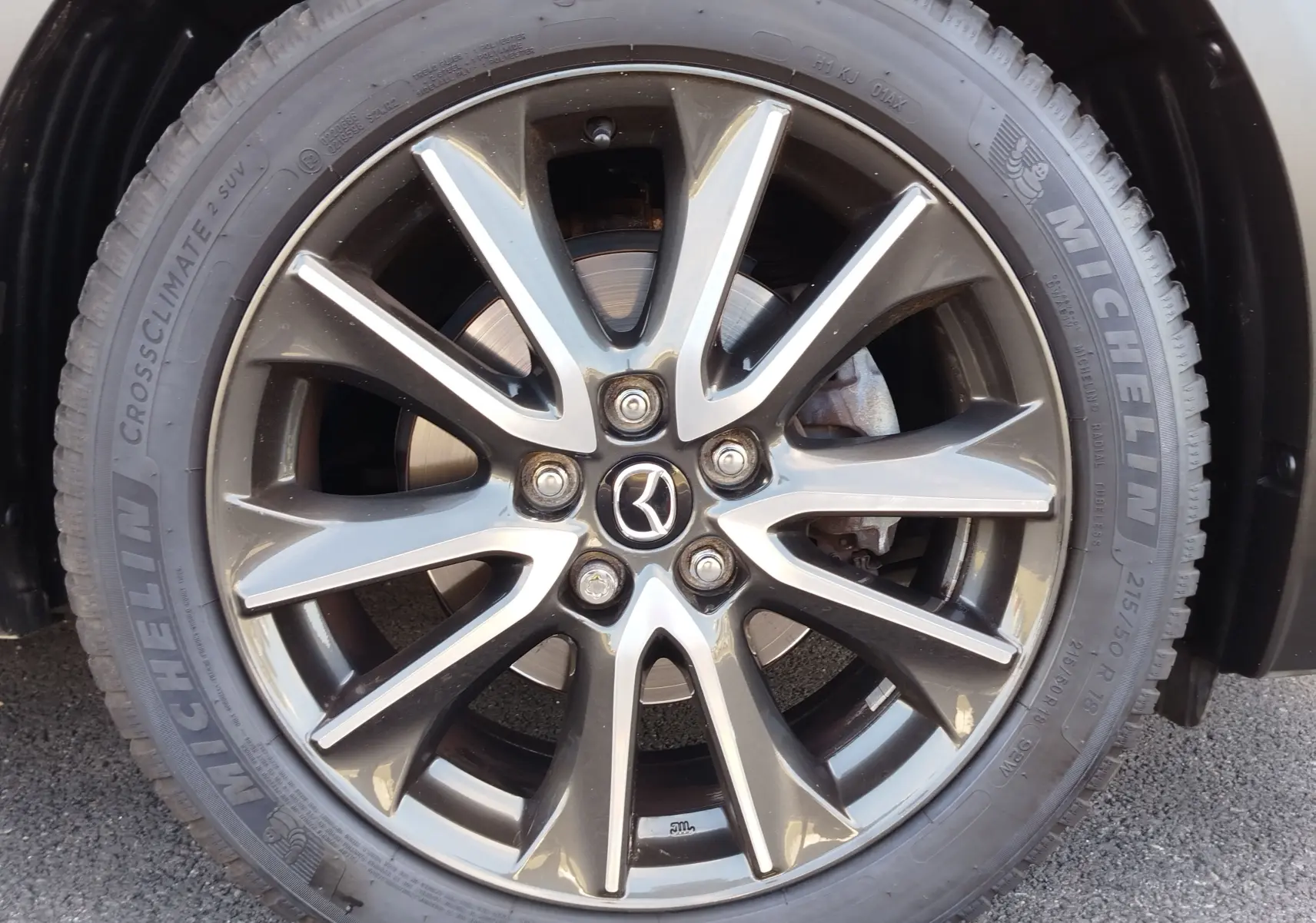 Gros plan sur la jante alliage gris foncé du Mazda CX-3 Dynamic Blue Mica, avec logo Mazda au centre et pneu Michelin visible.