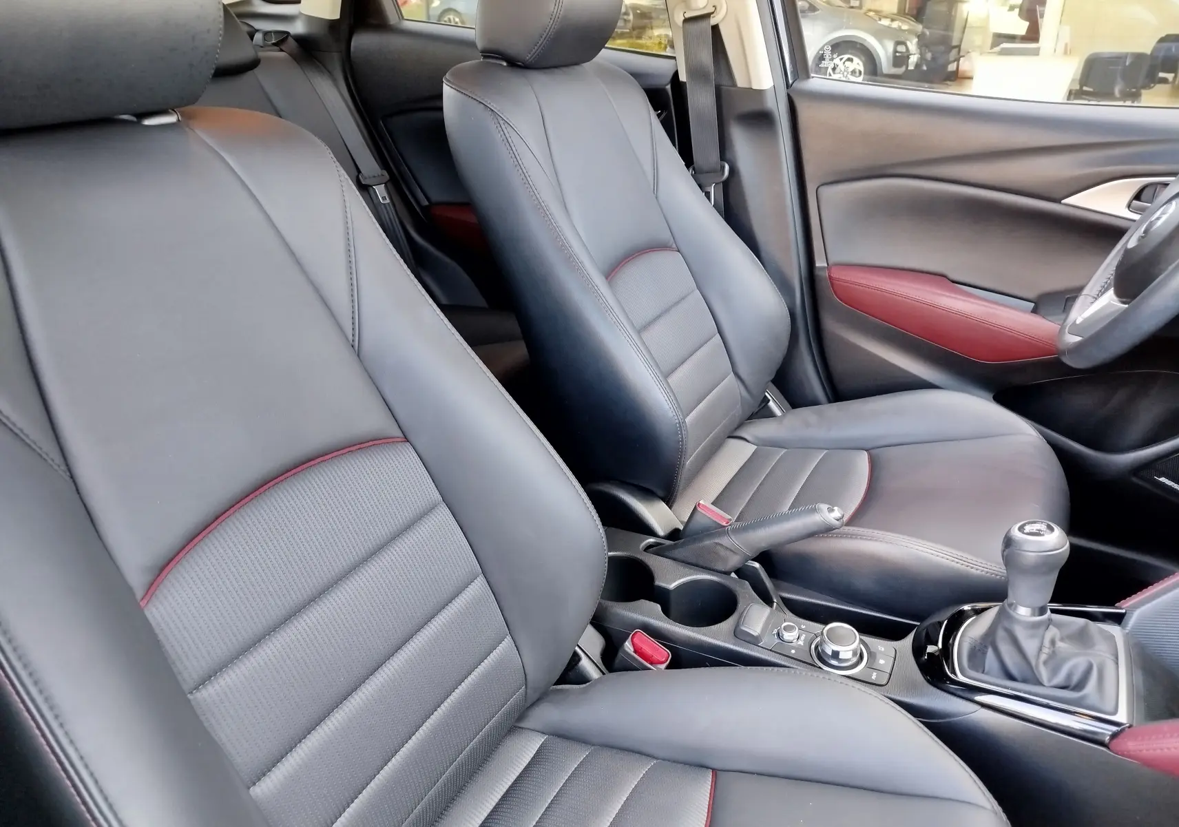 Vue intérieure côté conducteur du Mazda CX-3 2016, sièges cuir noir avec surpiqûres rouges et détails rouges sur les portes.