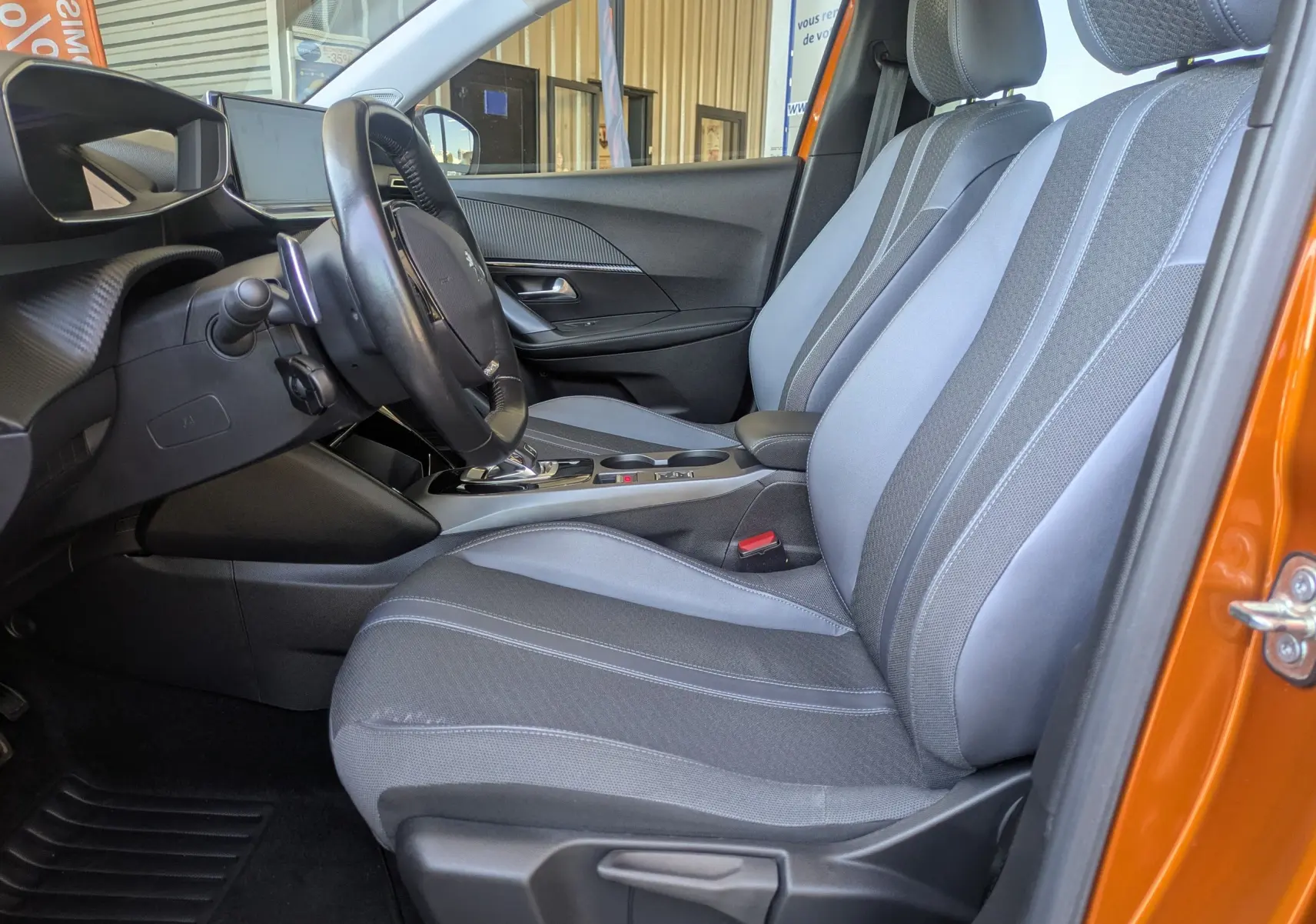 Vue intérieure côté conducteur de la Peugeot 2008 orange 2021, montrant les sièges gris et la console centrale avec boîte automatique.