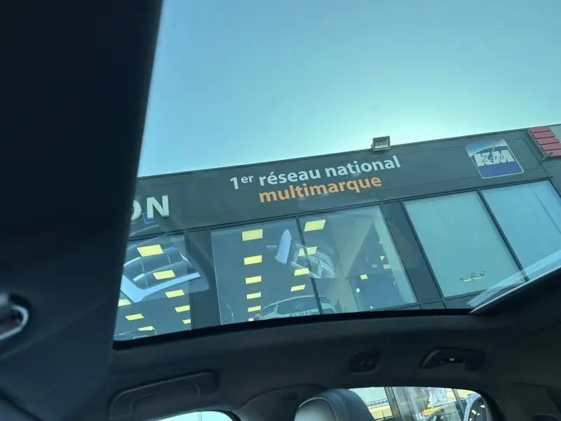 Vue depuis l'intérieur vers le toit ouvrant d'un Porsche Cayenne coupe noir métal, 3/4 avant droit visible sous un ciel clair.