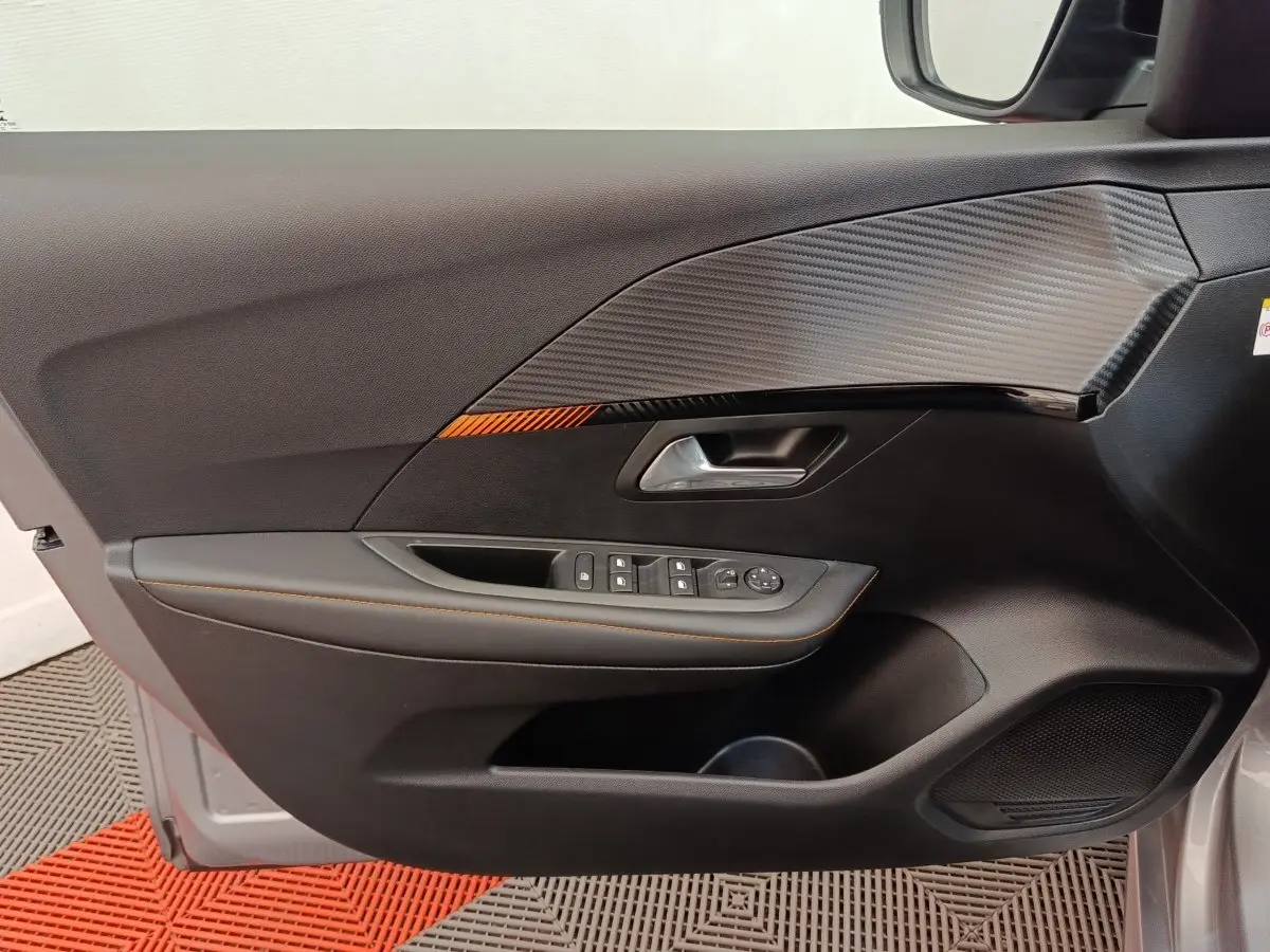 Vue intérieure côté gauche de la porte avant gris clair d’une Peugeot 208 Electrique ROADTRIP avec insert décor carbone et surpiqûres orange.