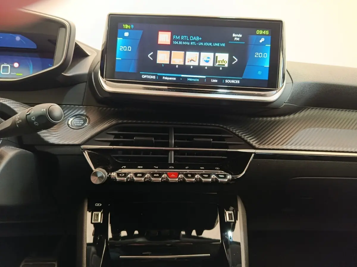 Vue intérieure centrée sur le tableau de bord noir avec écran tactile 10 pouces et commandes piano du Peugeot 208 électrique gris clair.