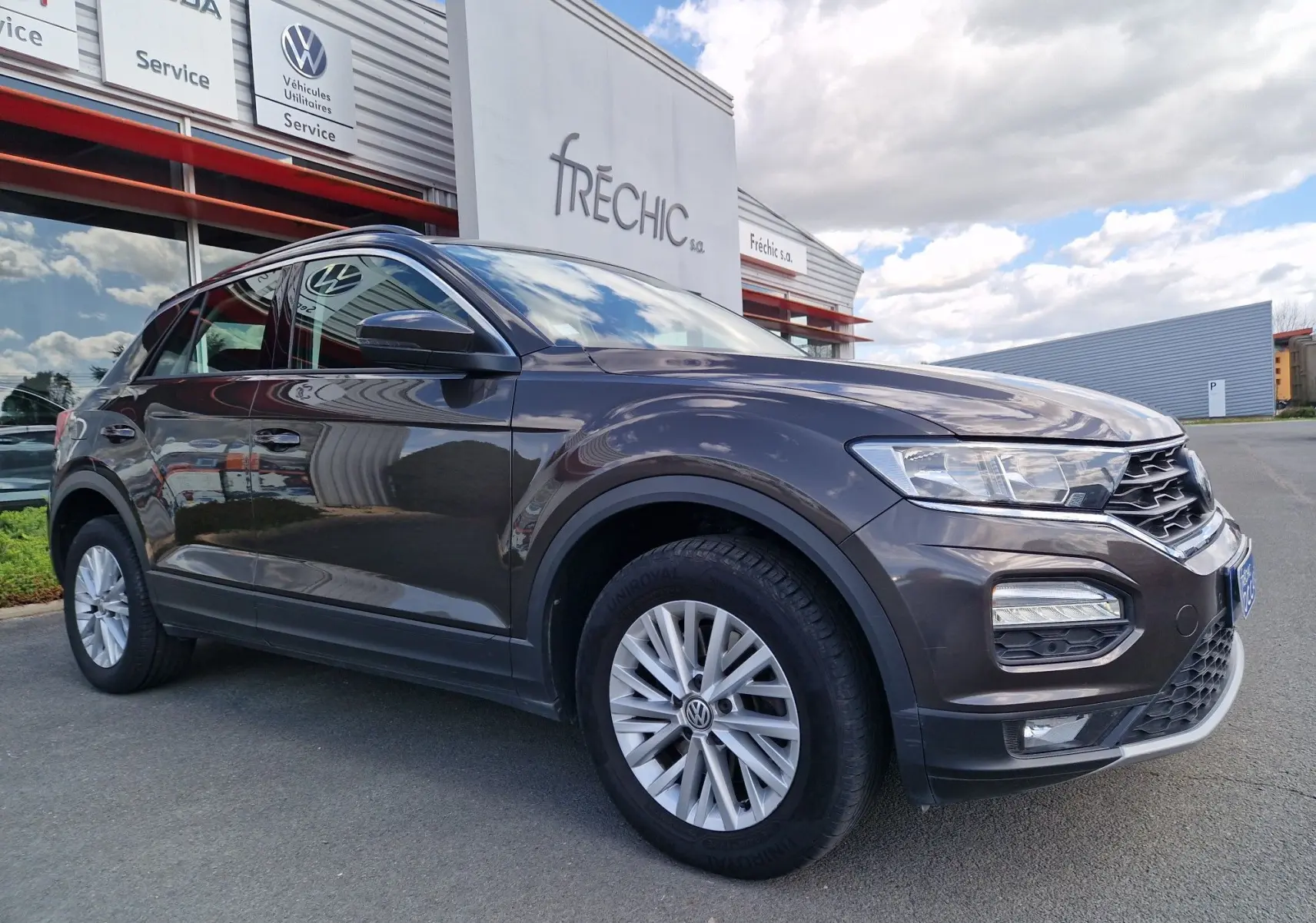 Volkswagen T-Roc chêne noir en 3/4 avant droit, avec jantes alliage et calandre distinctive sous un ciel nuageux.