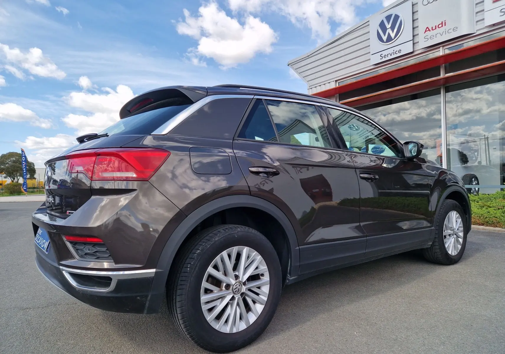 Vue 3/4 arrière droite d'un Volkswagen T-Roc chêne noir stationné devant un service Volkswagen avec jantes alliage visibles.