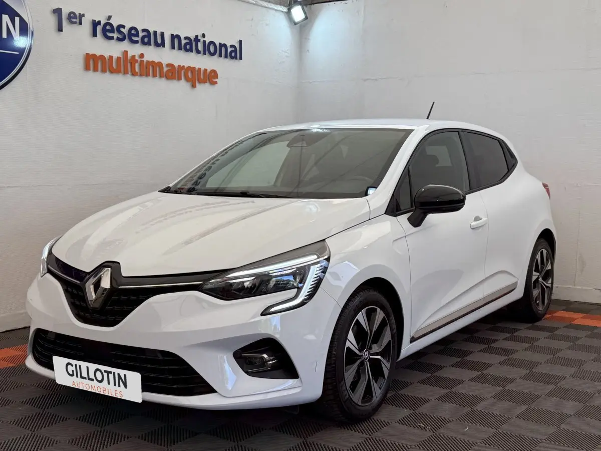 Renault Clio blanc vue 3/4 avant droit, avec phares LED et jantes alliage noires, en intérieur showroom.