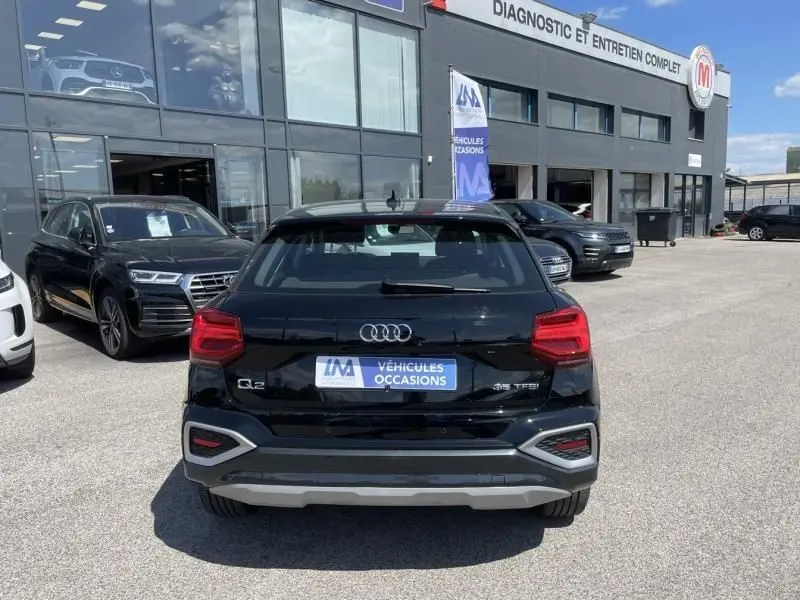 Vue arrière d'un Audi Q2 noir métal 2022 TFSI 150CV avec logo et feux arrière distinctifs en stationnement devant un garage.