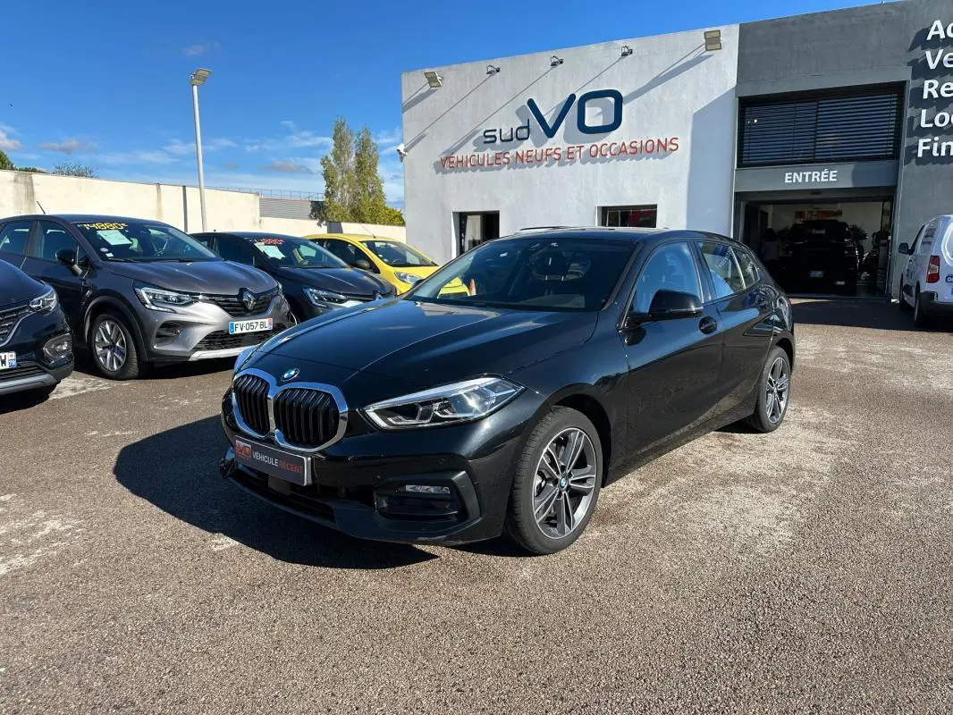 BMW Série 1 F40 118d noire vue en 3/4 avant droit sur parking devant concession Sud VO.