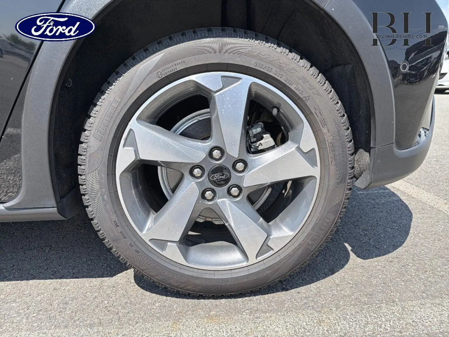 Gros plan sur la roue avant gauche d'une Ford Focus noire métallisée, jante alliage 5 branches avec logo Ford central.