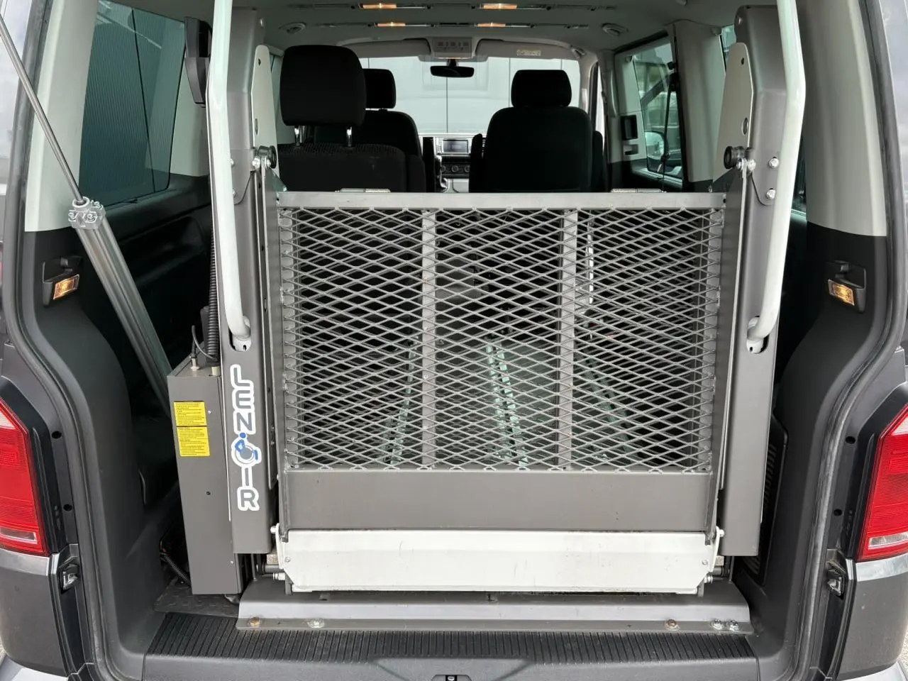 Vue arrière ouverte du Volkswagen Multivan T6 gris Indium avec rampe d'accès TPMR déployée dans le coffre.