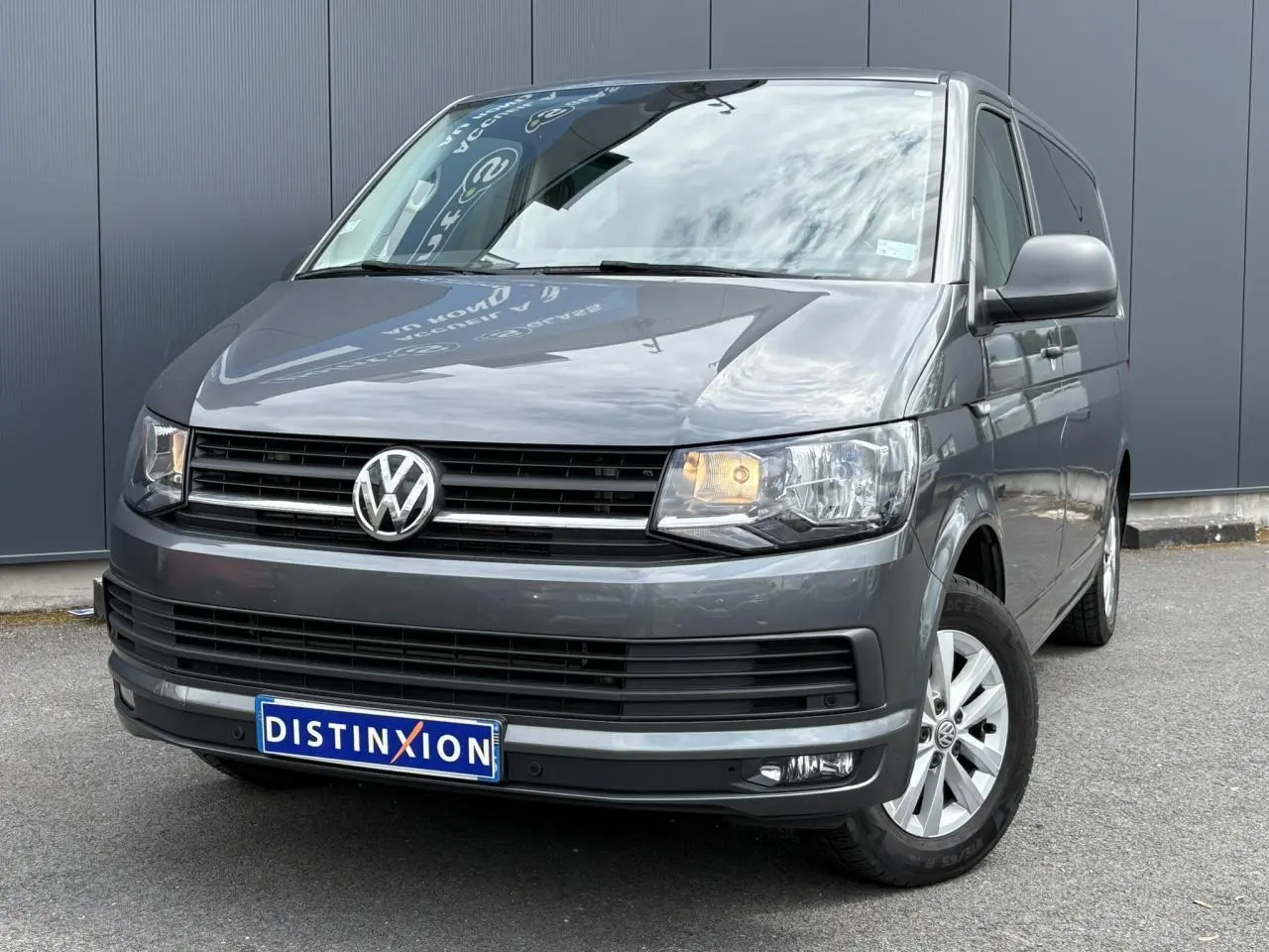 Volkswagen Multivan T6 gris Indium vu en 3/4 avant droit avec phares allumés et jantes alliage.