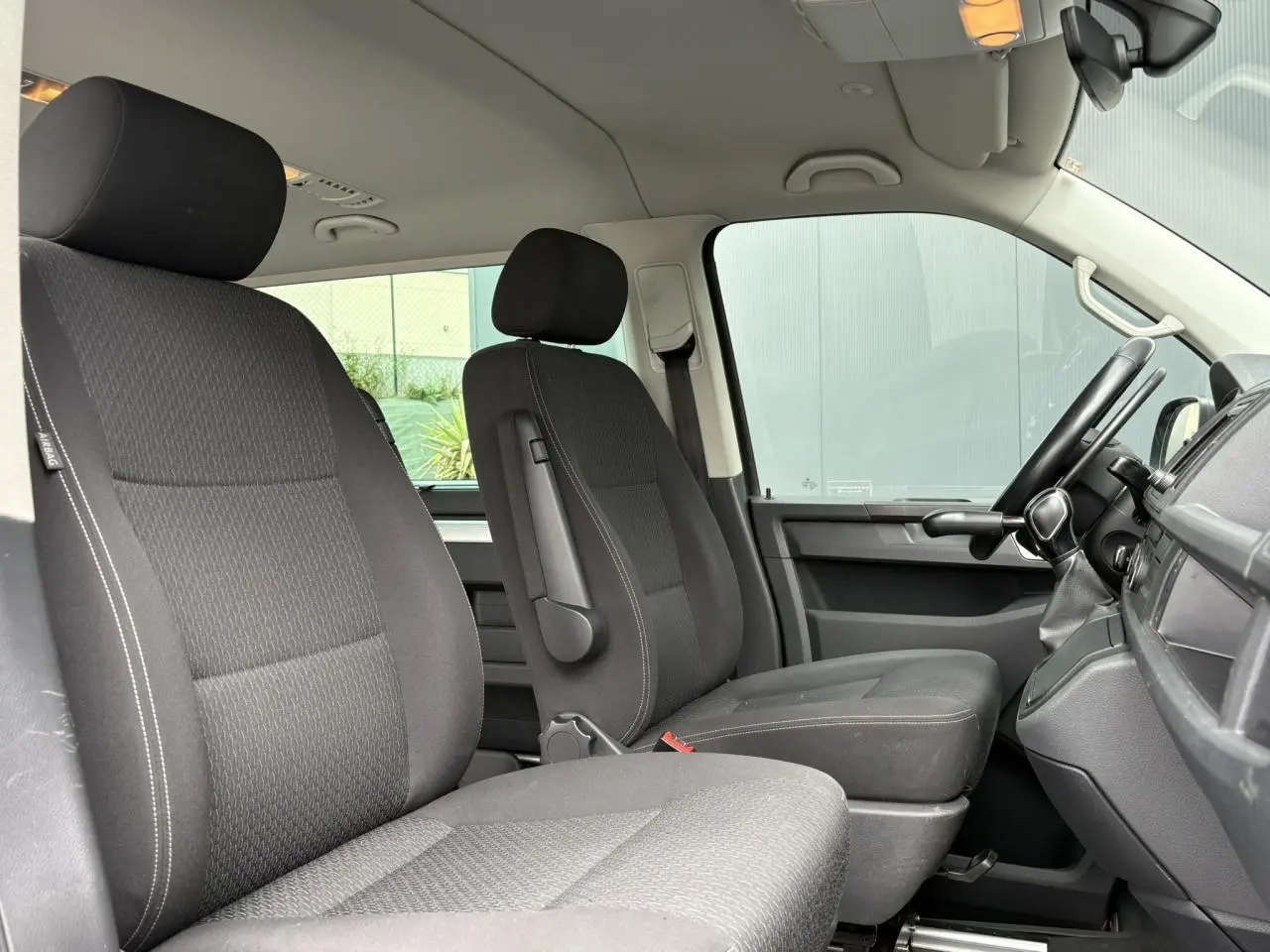 Vue intérieure côté conducteur du Volkswagen Multivan T6 gris Indium montrant les sièges avant en tissu noir et le tableau de bord.