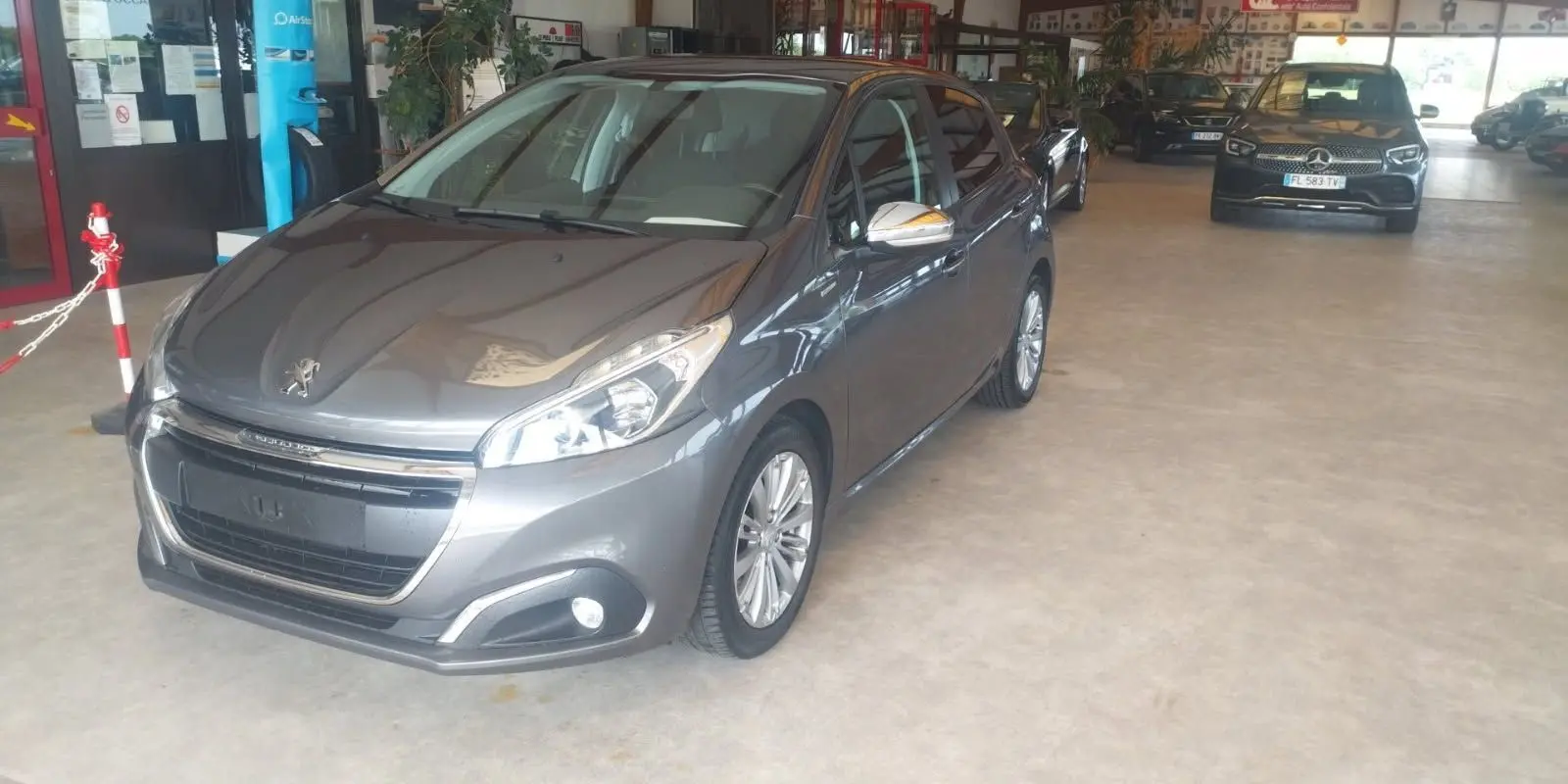 Peugeot 208 gris métal vue 3/4 avant droit dans un showroom avec plusieurs autres véhicules en arrière-plan.