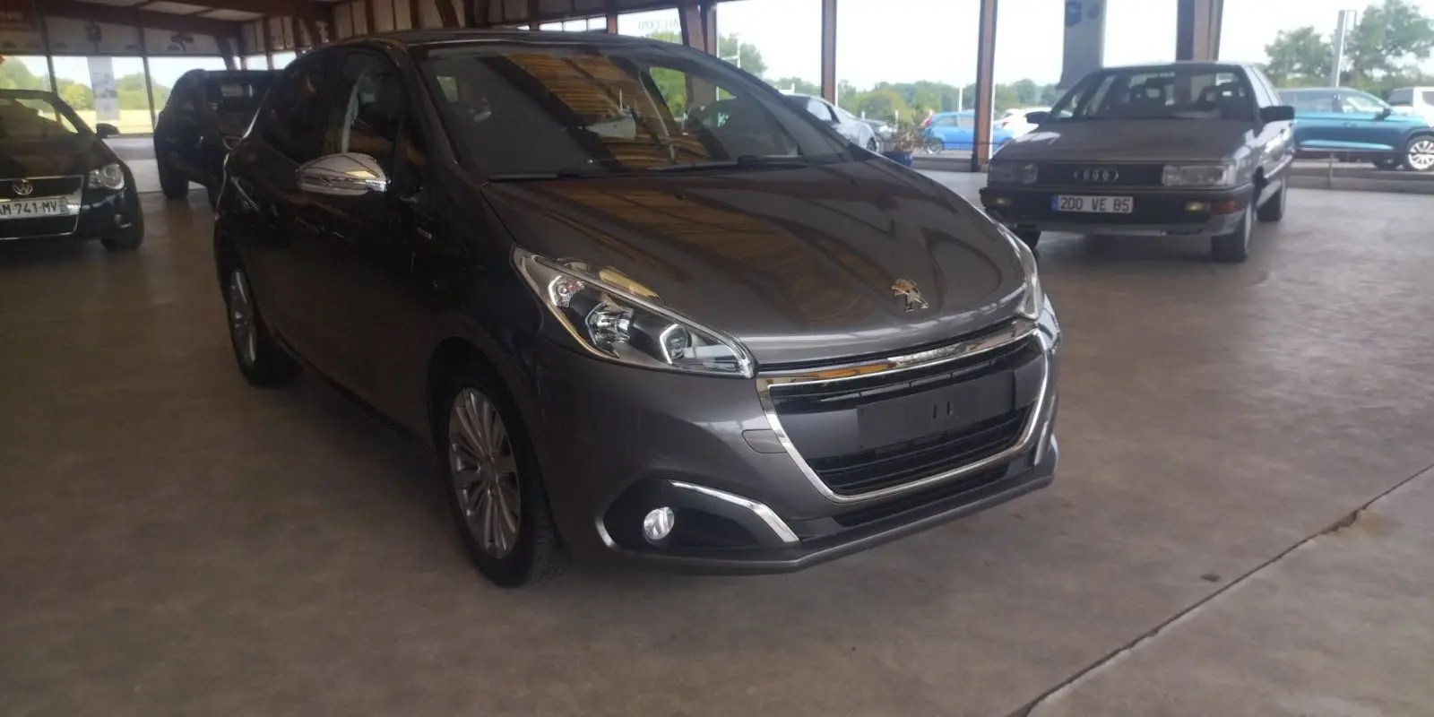 Peugeot 208 gris métal vue 3/4 avant droit, avec calandre chromée et phares modernes, en intérieur d'exposition.