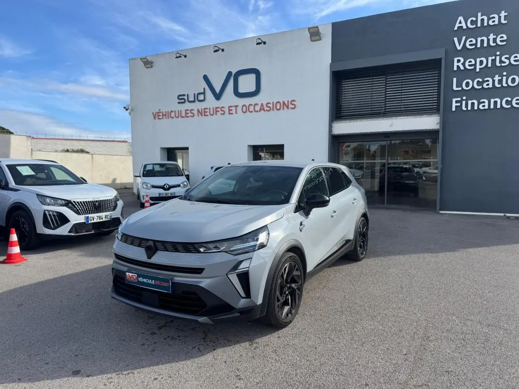 Renault Symbioz gris clair en 3/4 avant droit, jantes alliage noires et toit noir, devant un concessionnaire.