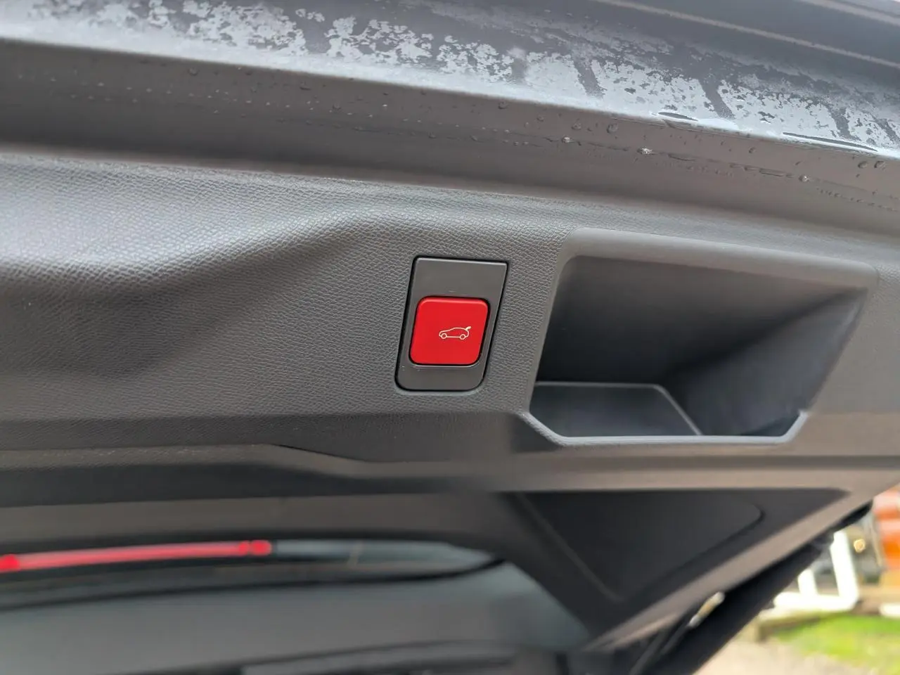 Bouton rouge d'ouverture électrique du hayon visible sous le coffre d'un Peugeot 5008 gris hybride 2025.