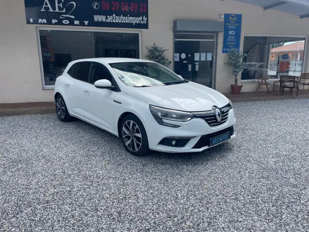 Renault Mégane IV blanc vue 3/4 avant droit, avec jantes alliage et calandre chromée, stationnée sur gravier devant un commerce.