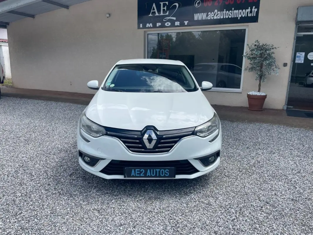 Vue frontale d'une Renault Mégane IV blanche, avec calandre chromée et phares LED, stationnée devant un garage.