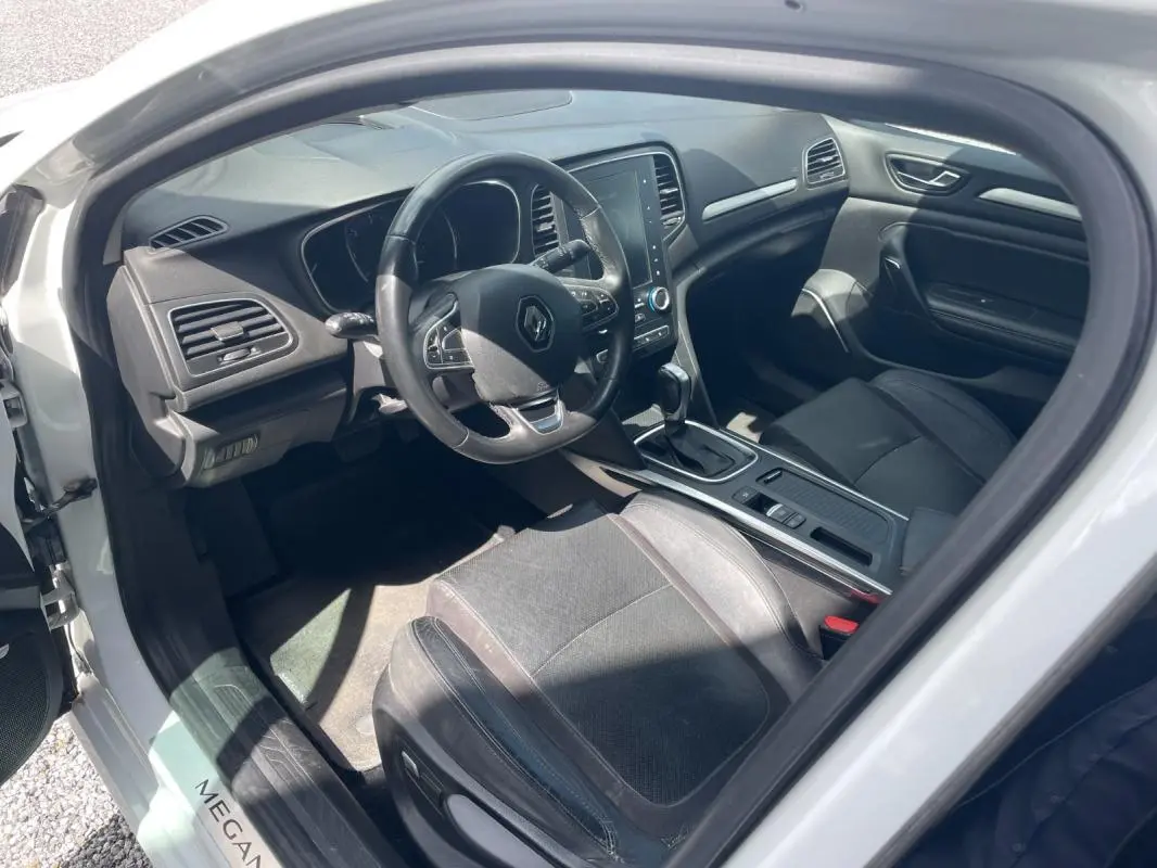 Vue intérieure côté conducteur de la Renault Mégane IV blanche de 2016, avec tableau de bord et console centrale visibles.