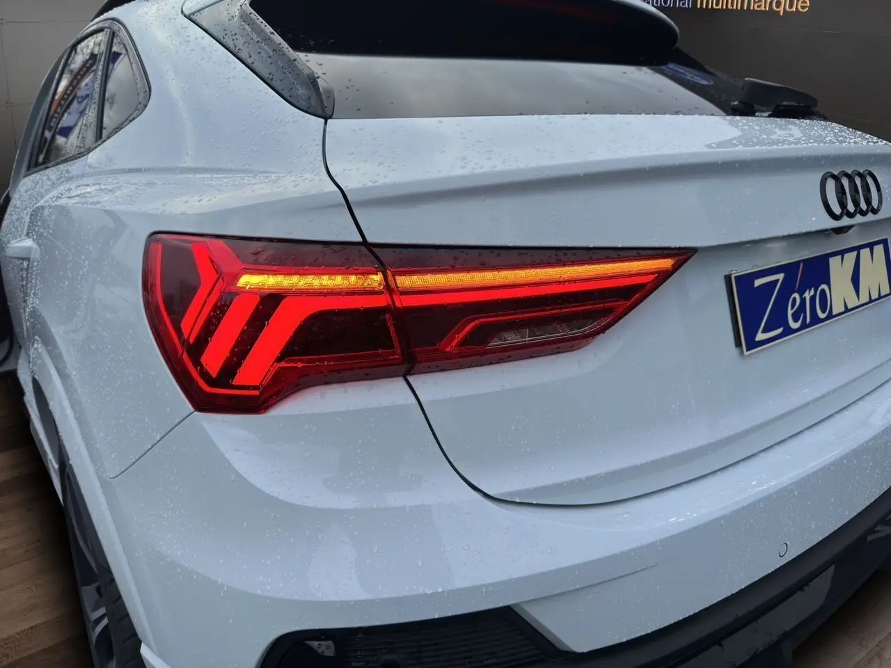 Vue 3/4 arrière droite d'un Audi Q3 Sportback blanc glacier avec feux arrière LED allumés et pluie sur la carrosserie.