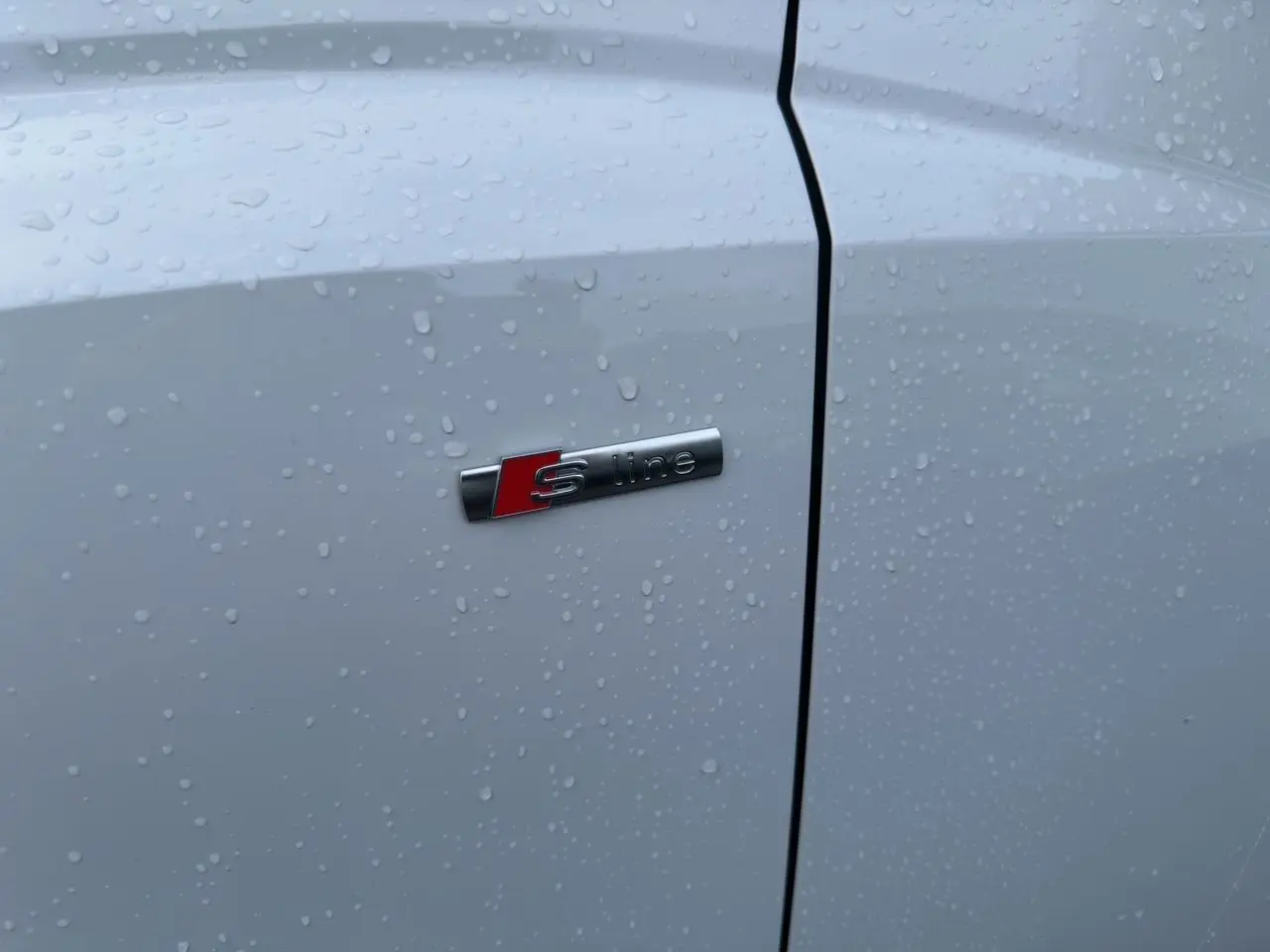 Gros plan sur l'emblème S line sur la porte blanche d'un Audi Q3 Sportback 2024 avec gouttes de pluie.