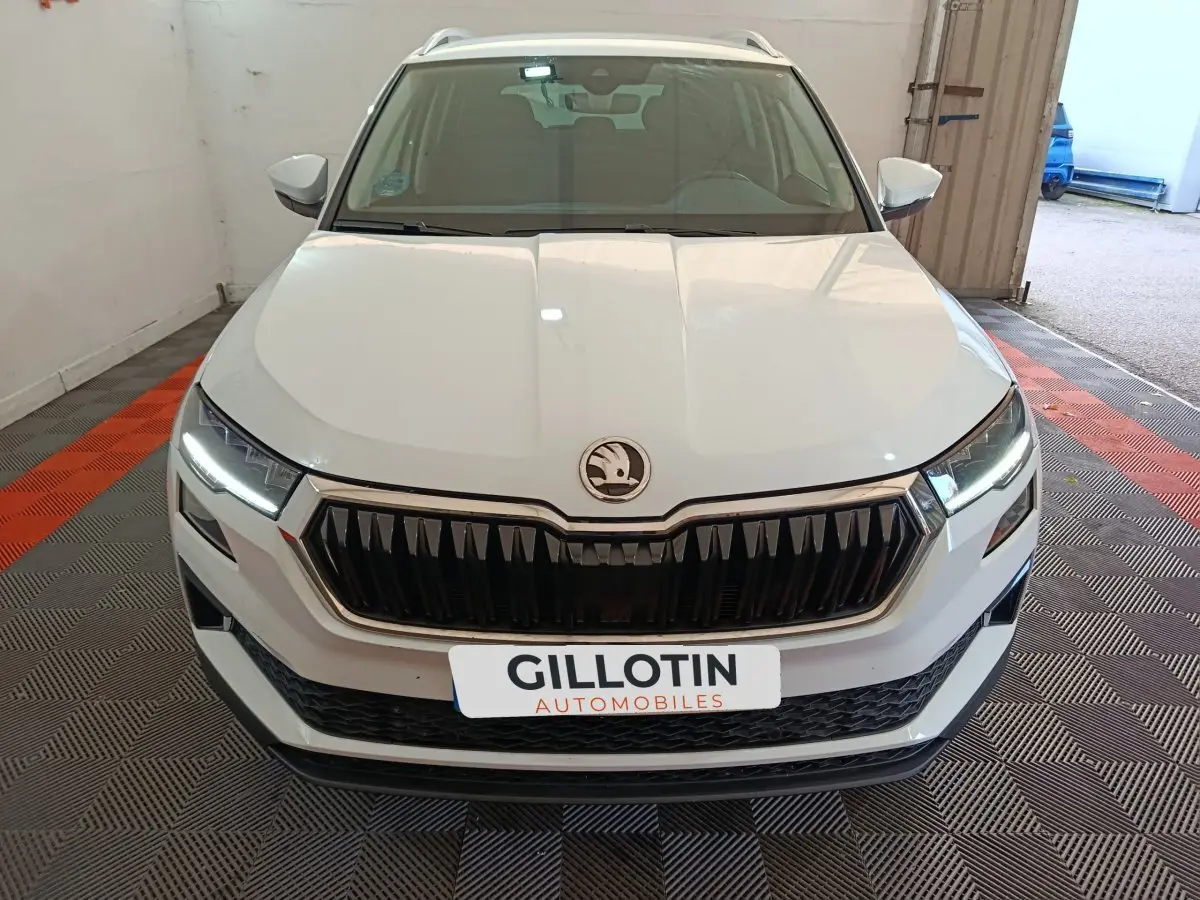 Vue avant d'un Skoda Karoq blanc 2024 avec calandre noire et phares LED allumés dans un garage.