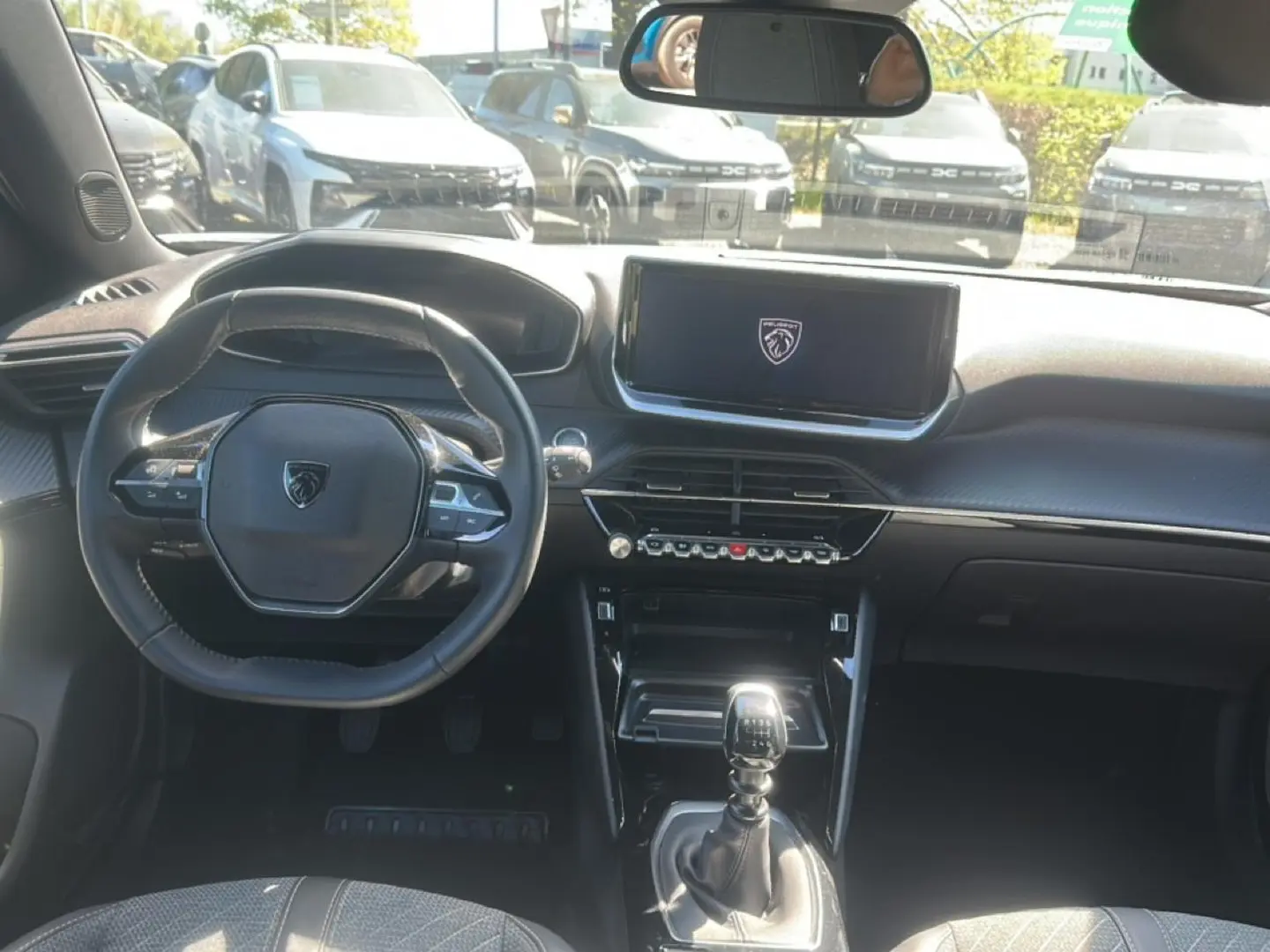 Vue intérieure avant du Peugeot 2008 gris Artense 2025, volant multifonction et écran tactile central allumé au logo Peugeot.