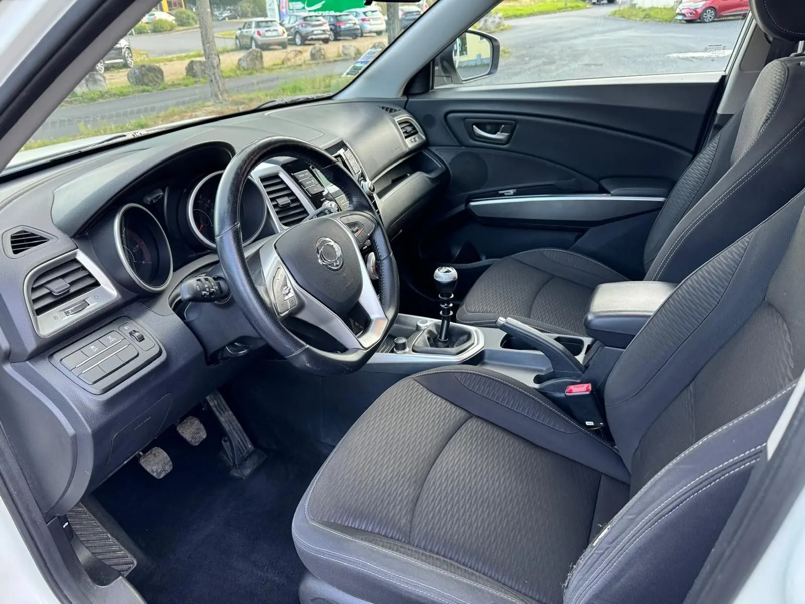 Intérieur côté conducteur du SsangYong Tivoli blanc 2016, volant cuir, levier de vitesse manuel et sièges tissu noirs.