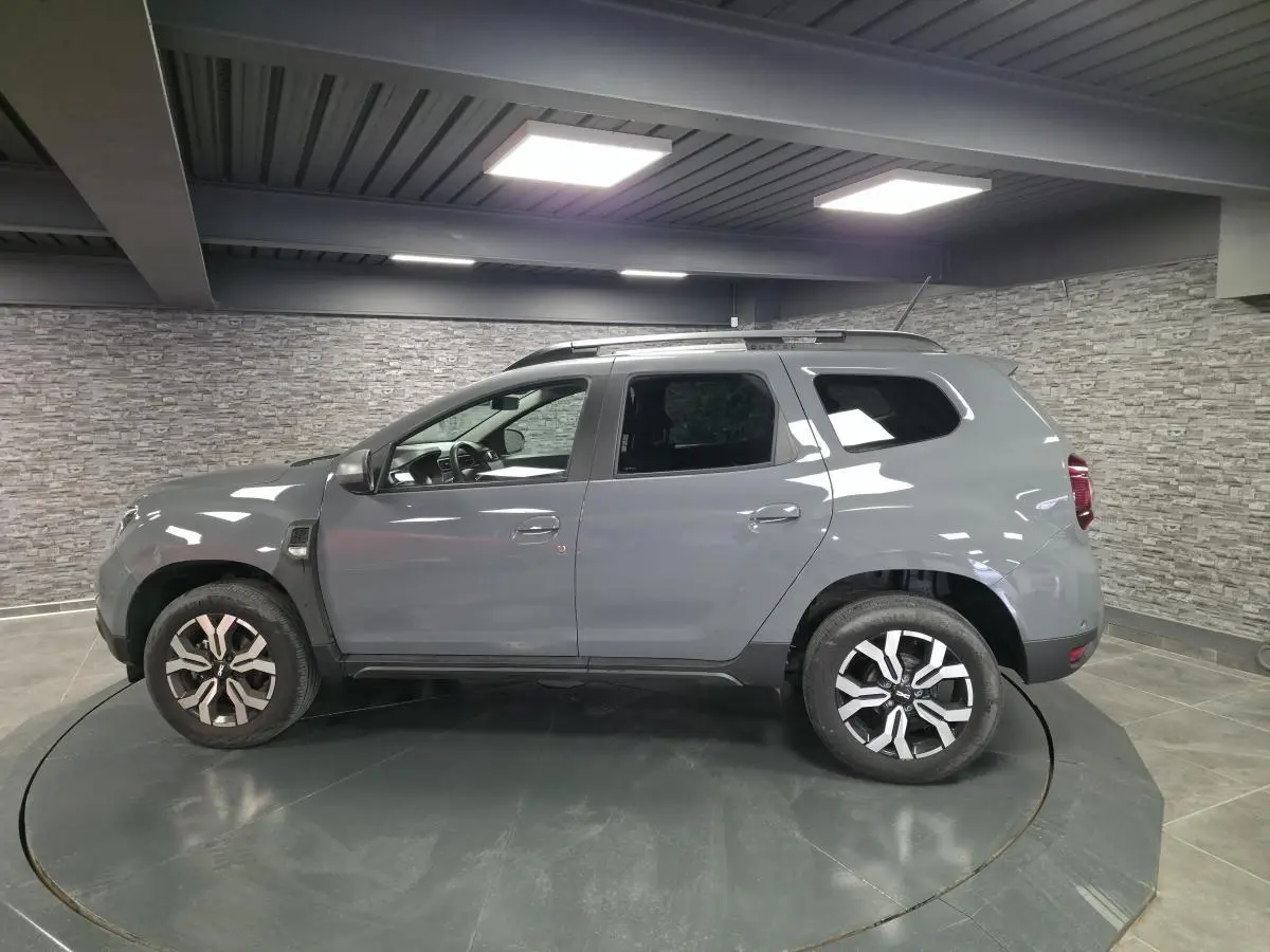 Vue de profil côté gauche d'un Dacia Duster 2024 gris urbain avec jantes alliage 17 pouces et barres de toit.