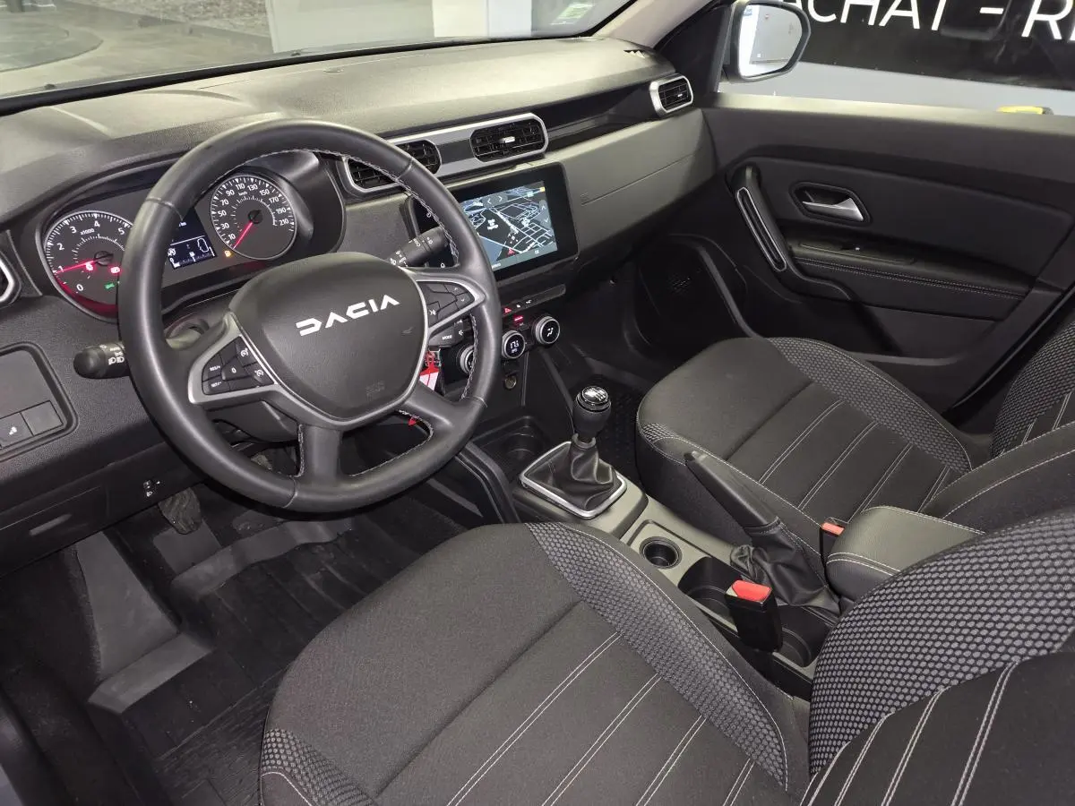 Vue intérieure avant droite du Dacia Duster 2024, tableau de bord noir avec écran tactile et volant multifonction.