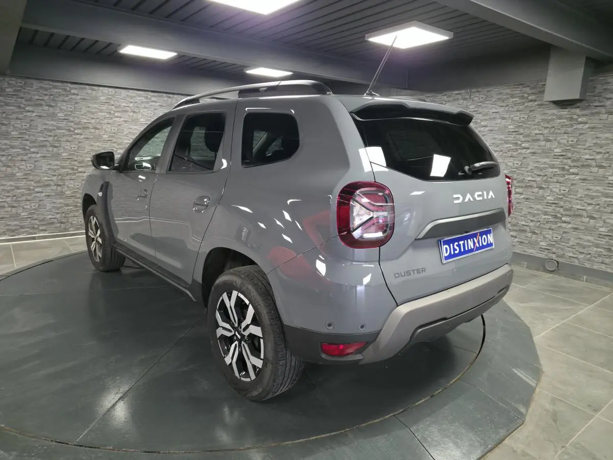 Vue 3/4 arrière droit du Dacia Duster 2024 gris urbain avec jantes alliage et barres de toit gris mégalithe.