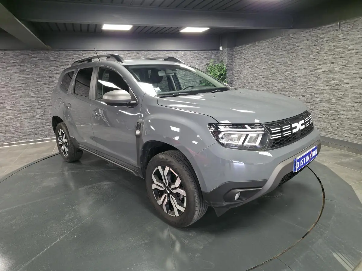 Vue 3/4 avant droit d'un Dacia Duster 2024 gris urbain avec jantes alliage et barres de toit gris mégalithe.